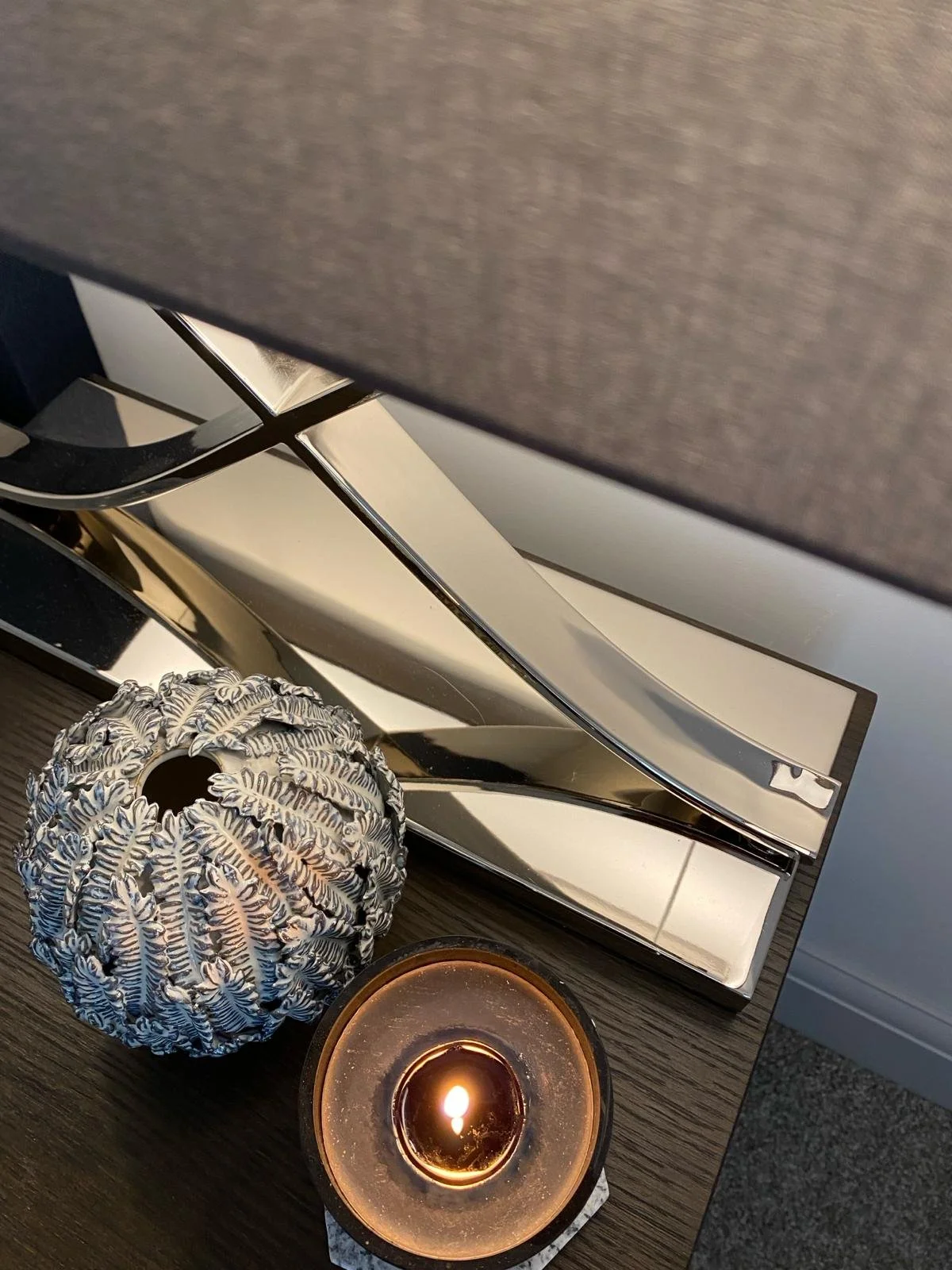 Decorative objects including a patterned spherical lamp, a lit candle, a shiny silver decorative piece, and part of a black-grey table with a wall or curtain in the background.