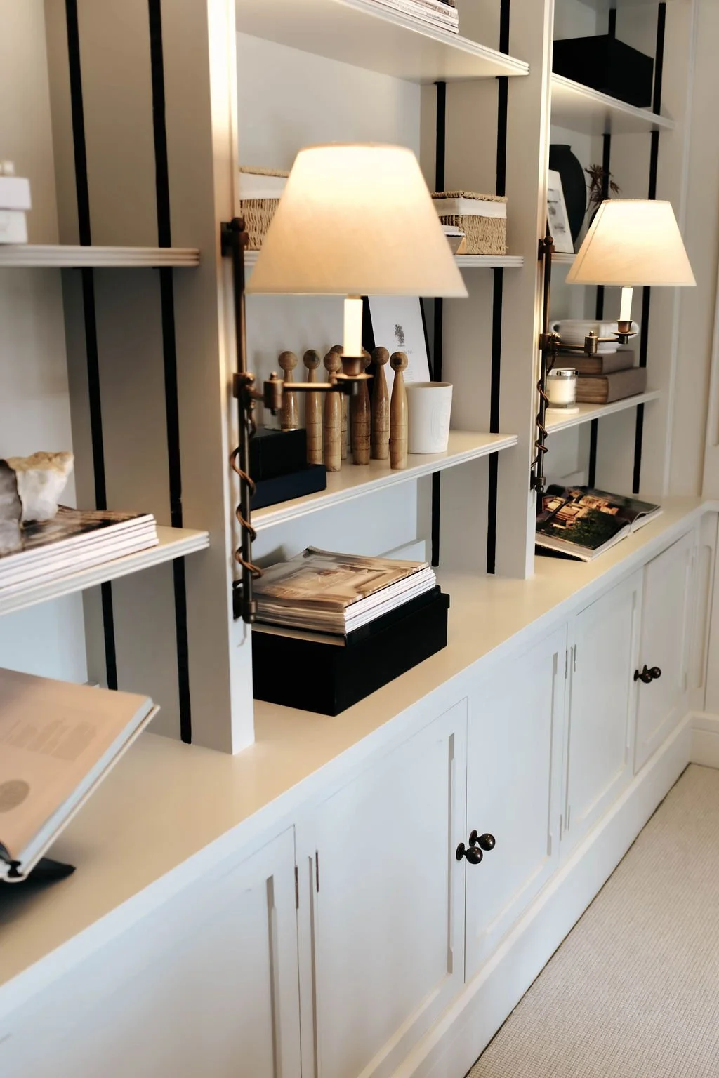 White bookcase with black accents, topped with decorative items like magazines, lamps, and vases, in a cozy, well-lit room.