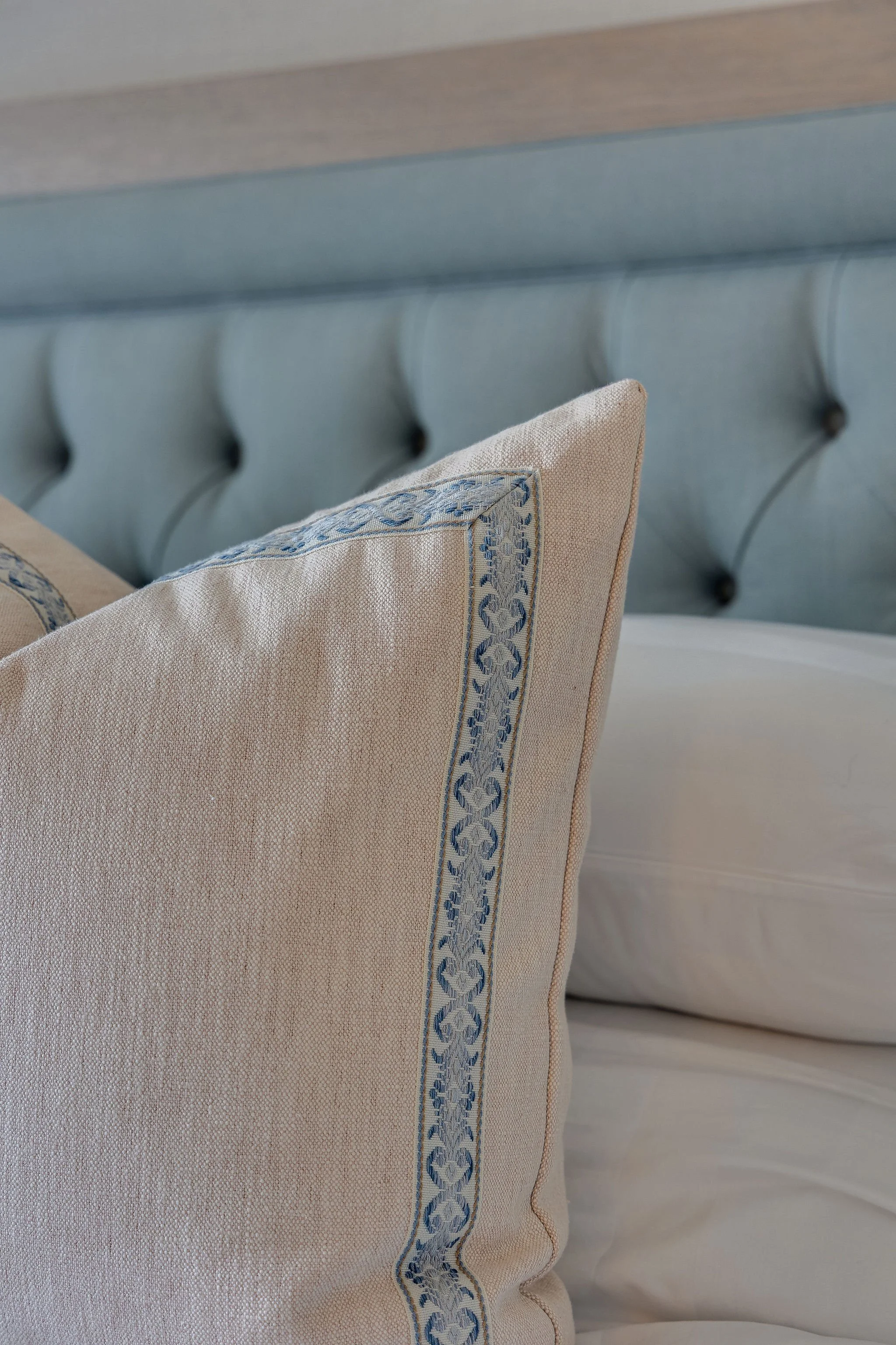 A close-up of a beige textured fabric pillow with a blue embroidered border, resting against a pale blue tufted headboard