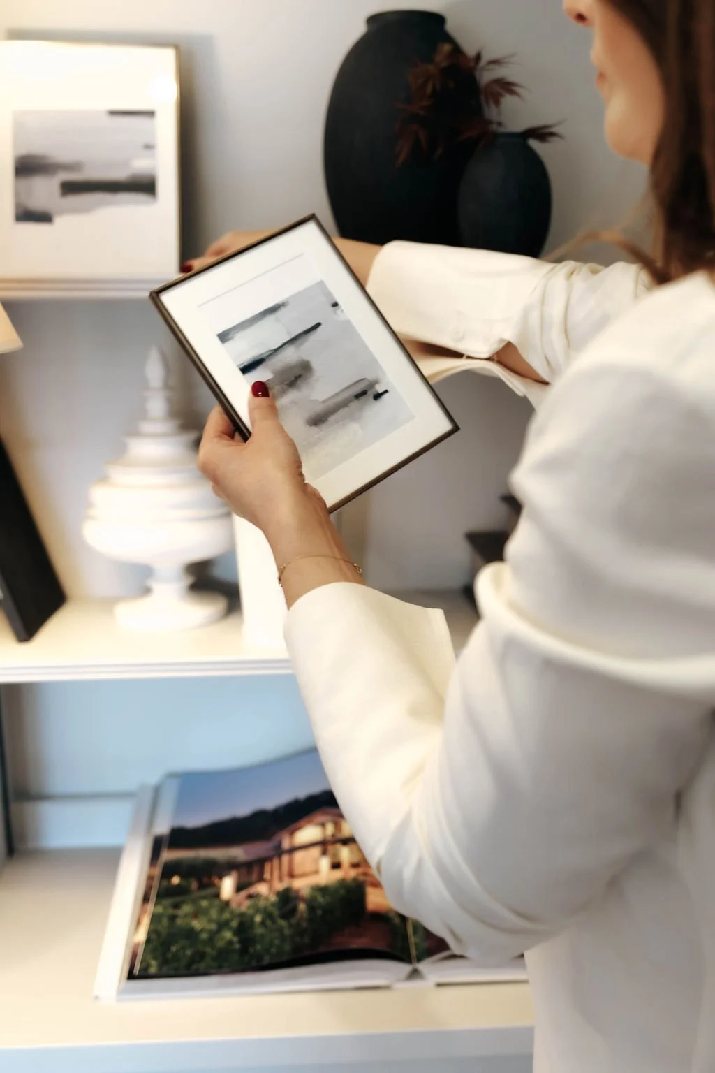 A woman in a white shirt is holding a framed photograph of a landscape with water and boats, while looking at a large open photo book on a shelf with images of buildings and nature.