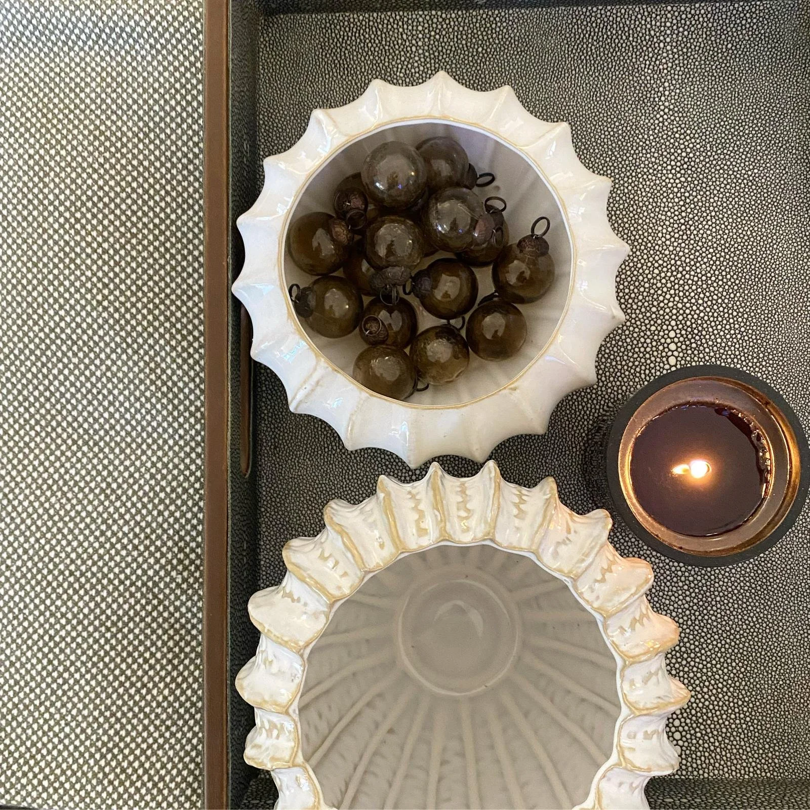 Decorative arrangement with dark Christmas ornaments in a white, scalloped ceramic bowl and a lit dark candle in a copper-colored holder on a textured black surface.
