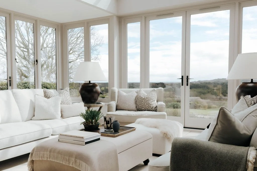 Bright living room with large windows showing a scenic outdoor landscape, white sofa, armchair, side tables with lamps, and decorative pillows.