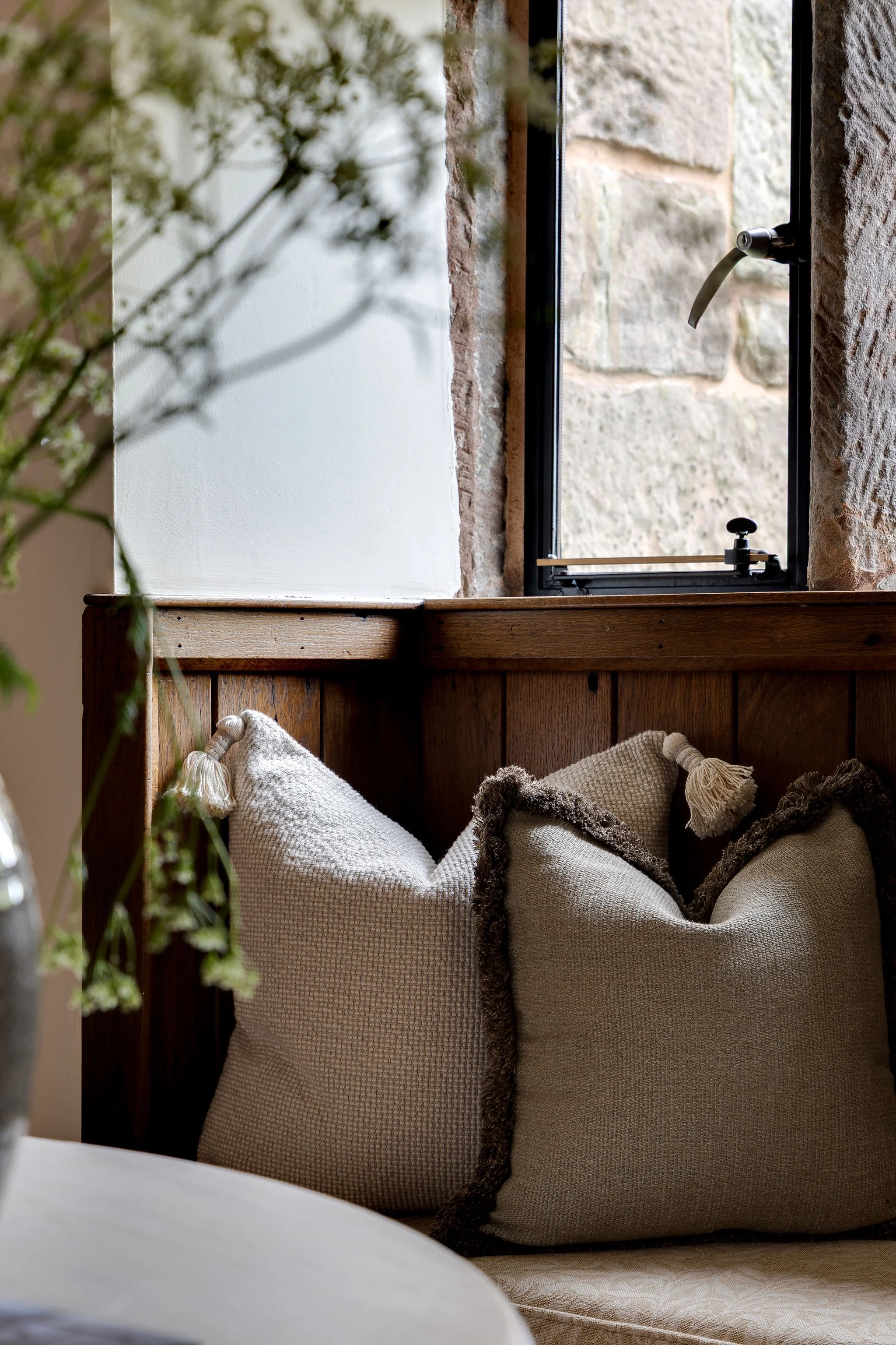 A cozy interior corner with a wooden headboard, beige and brown throw pillows, a window with a stone wall outside, and a blurred plant in the foreground.
