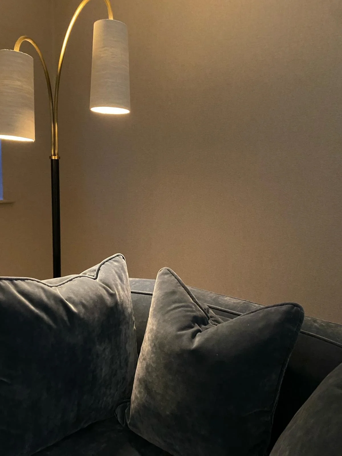 Living room with a gray sofa and pillows, beige wall, and a tall floor lamp with three cylindrical lampshades emitting warm light.