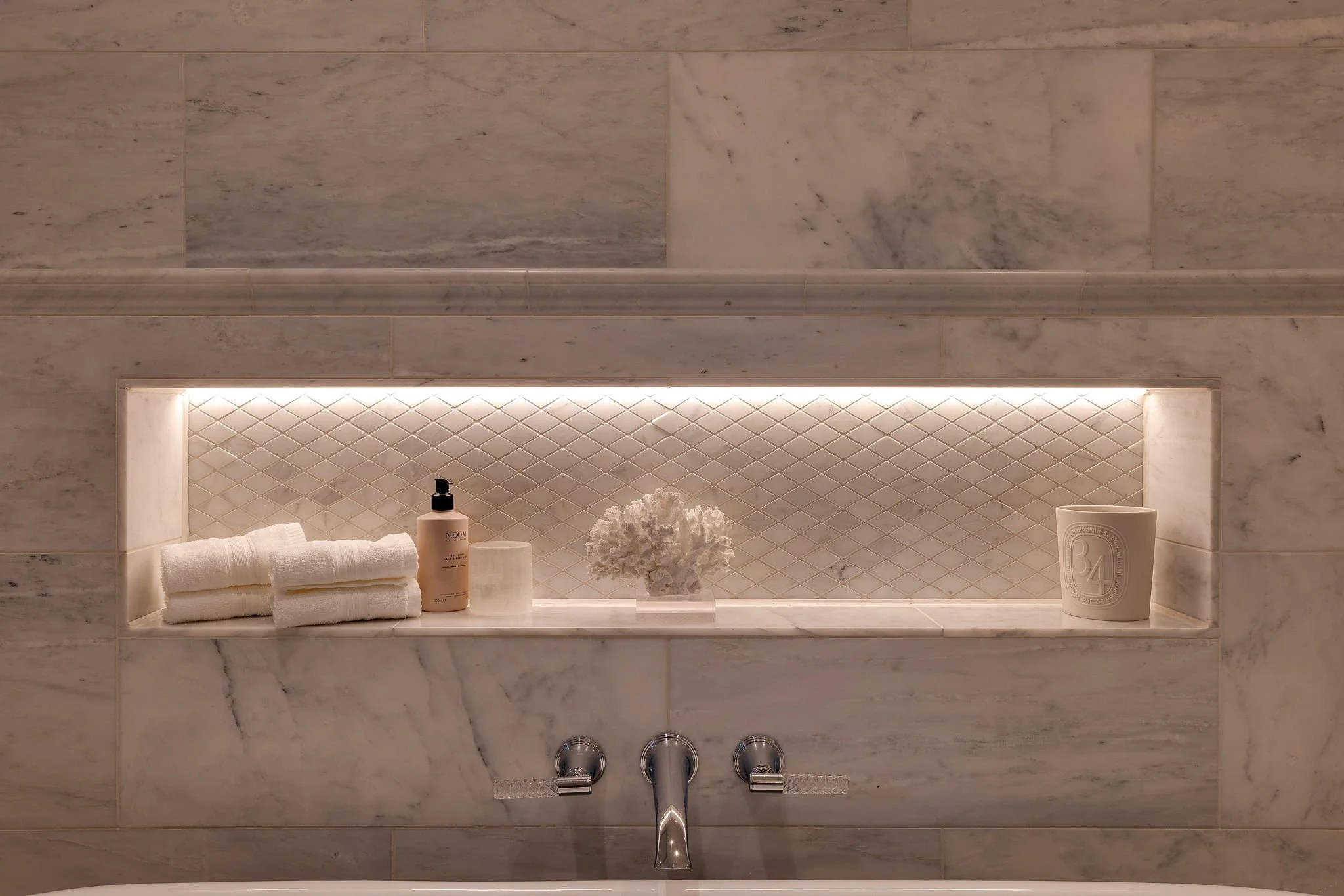 Bathroom shelf with towels, a soap dispenser, a decorative coral piece, a cup, and a bottle of skincare product against a marble wall, illuminated by built-in lighting.