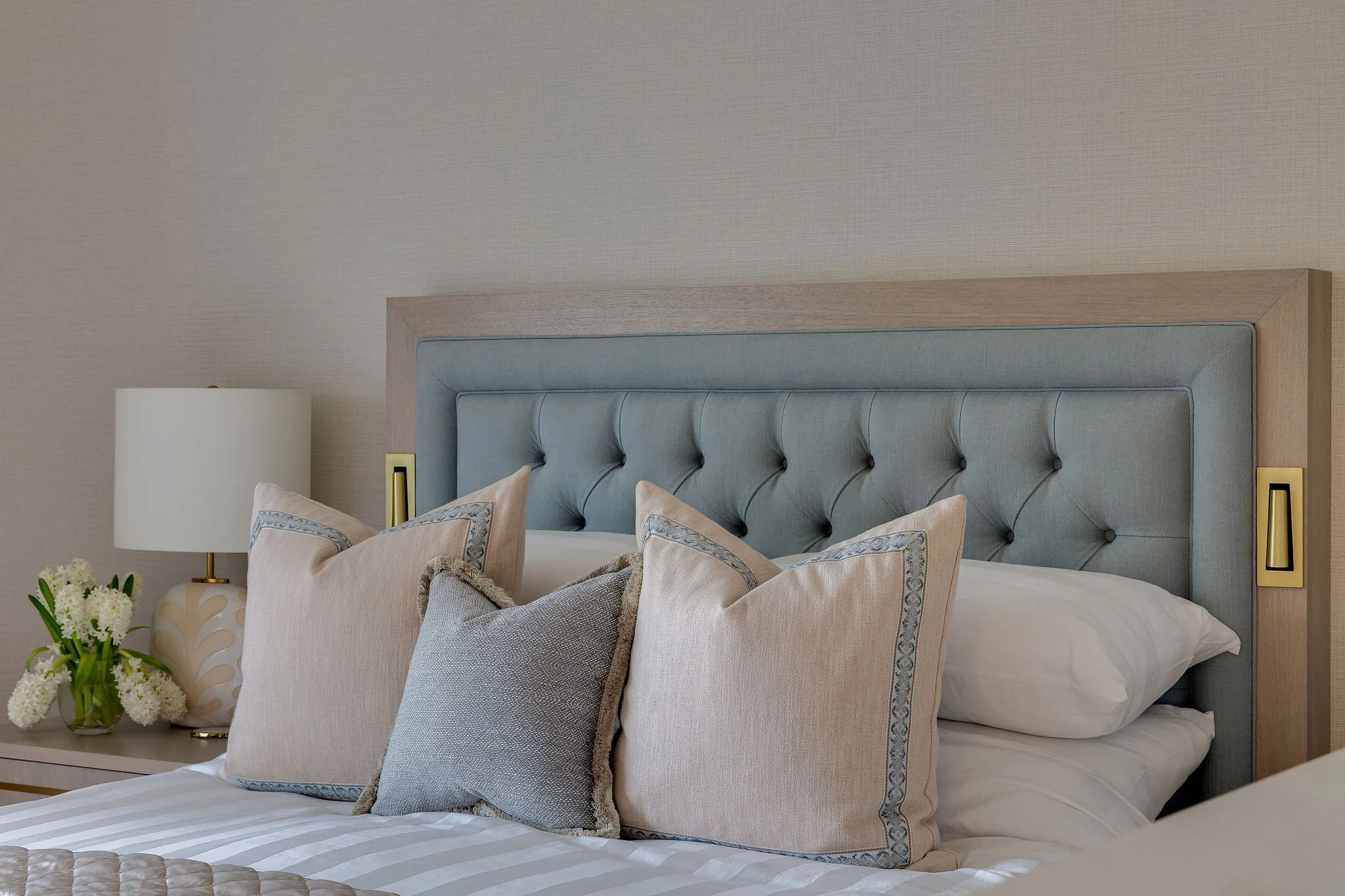 A bedroom with a large upholstered headboard, multiple throw pillows, a bedside table with a vase of white flowers, and a lamp, with beige walls and modern decor.