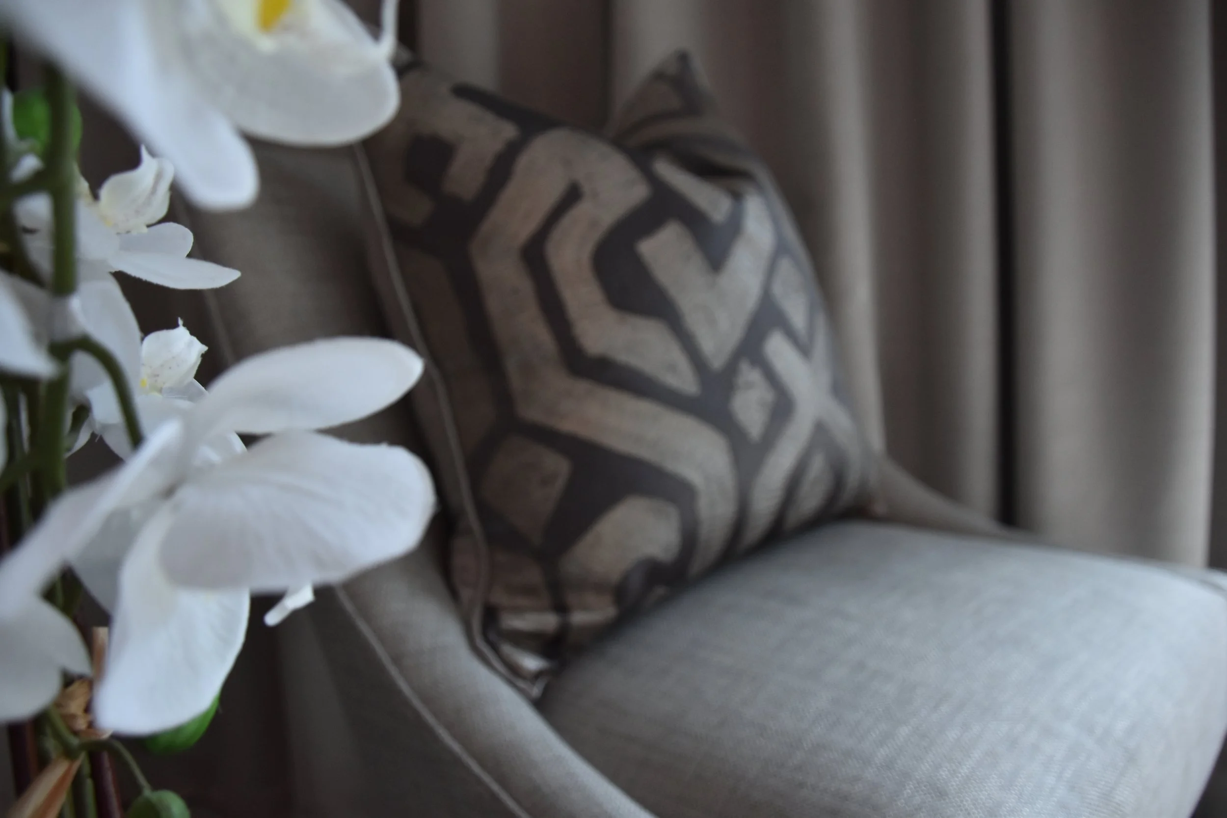 Close-up of a patterned decorative pillow on a couch, partially covered by white orchid flowers and beige curtains in the background.