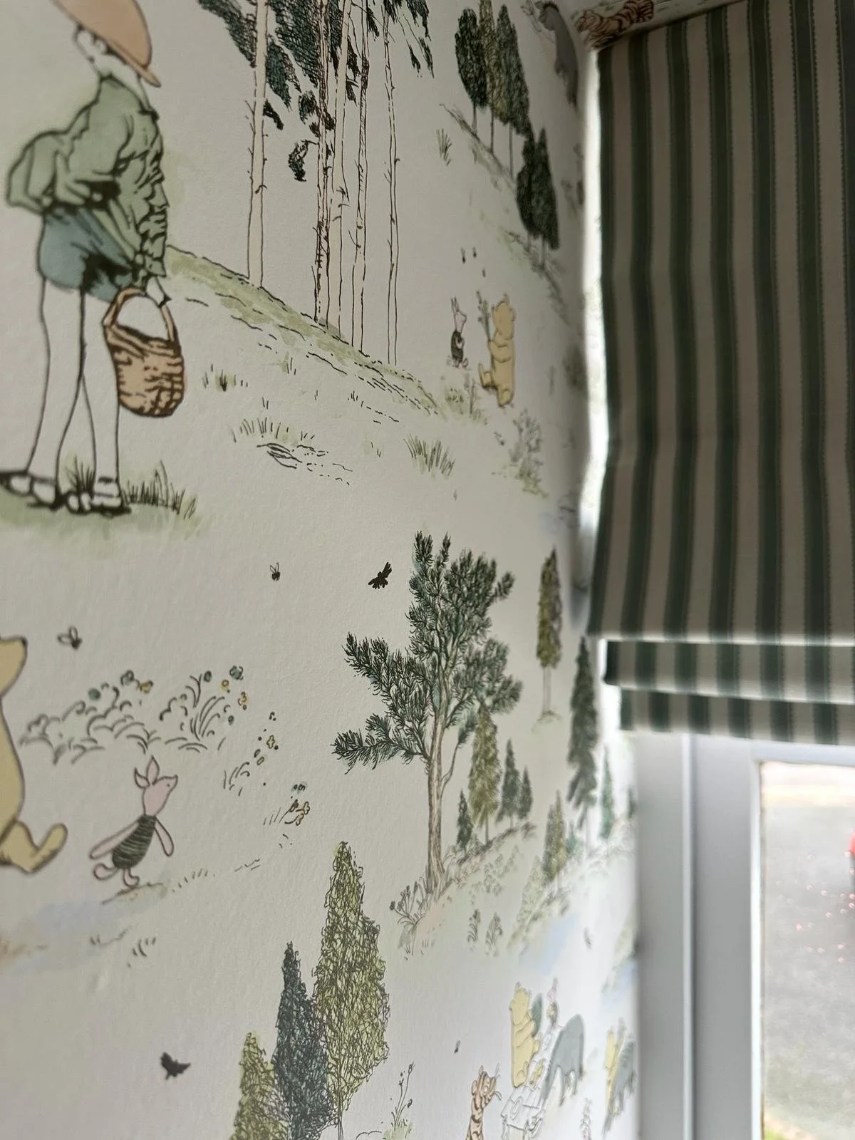 Close-up view of a fabric with a illustrated forest scene, including trees, animals like a rabbit and a bear, and a person holding a basket, with a window with striped curtains in the background.