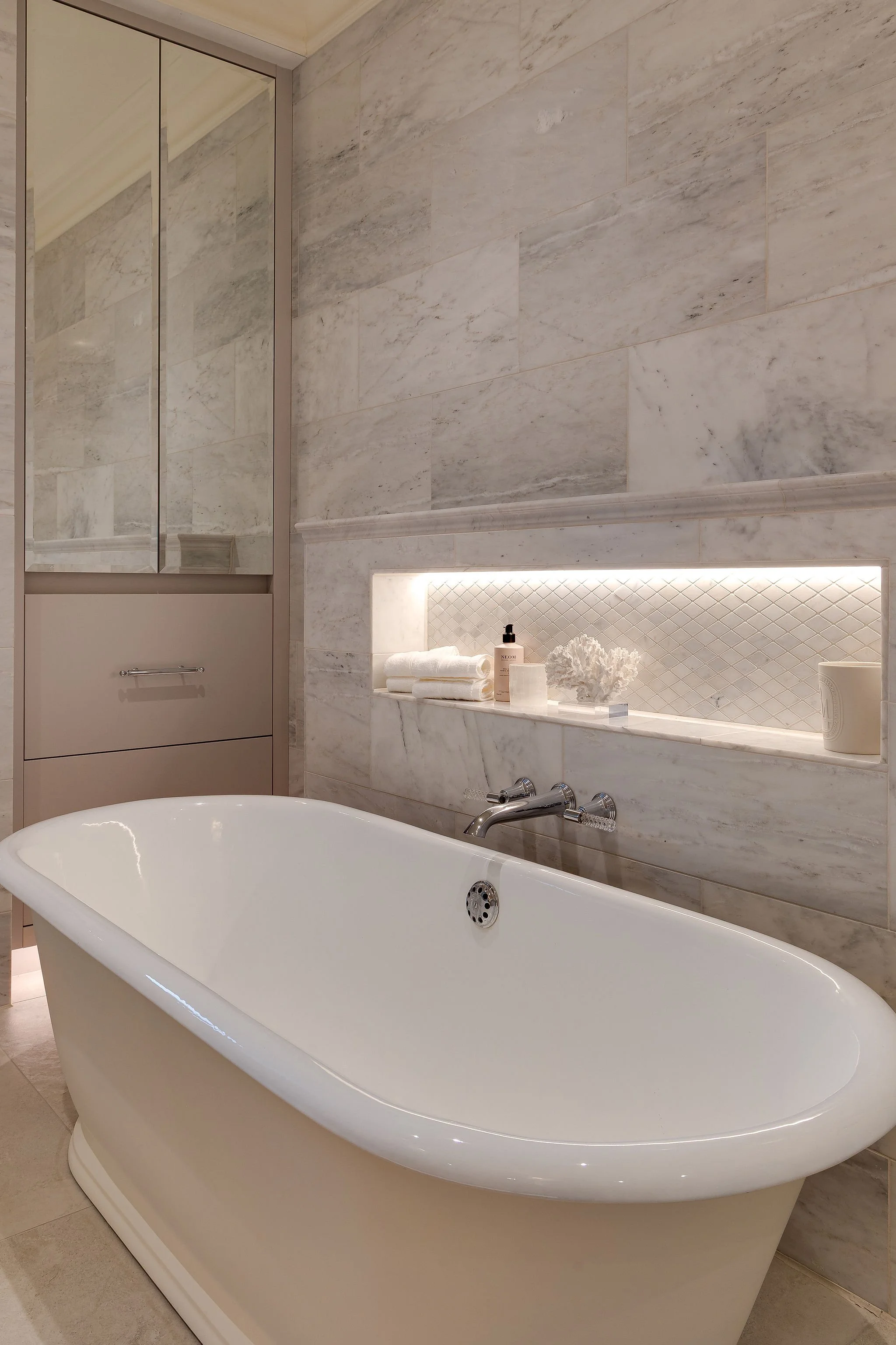 A bathroom with a white freestanding bathtub, marble wall tiles, a built-in shelf with white towels, a bottle of soap, decorative paper flowers, and a mirrored cabinet.
