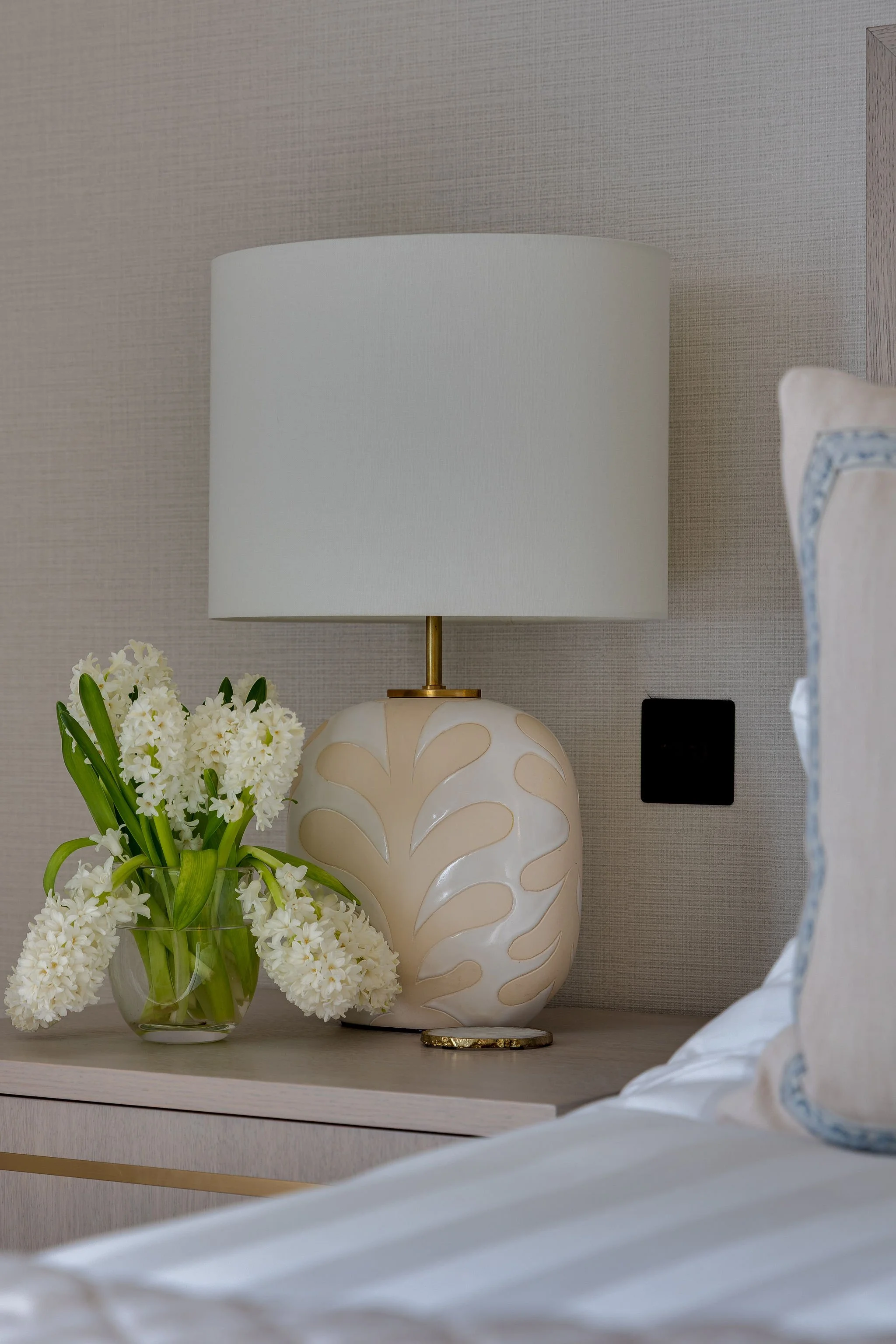 A bedside table with a white table lamp, a vase of white flowers, and a ceramic decorative object, set against a textured beige wall with a bed partially visible in the foreground.