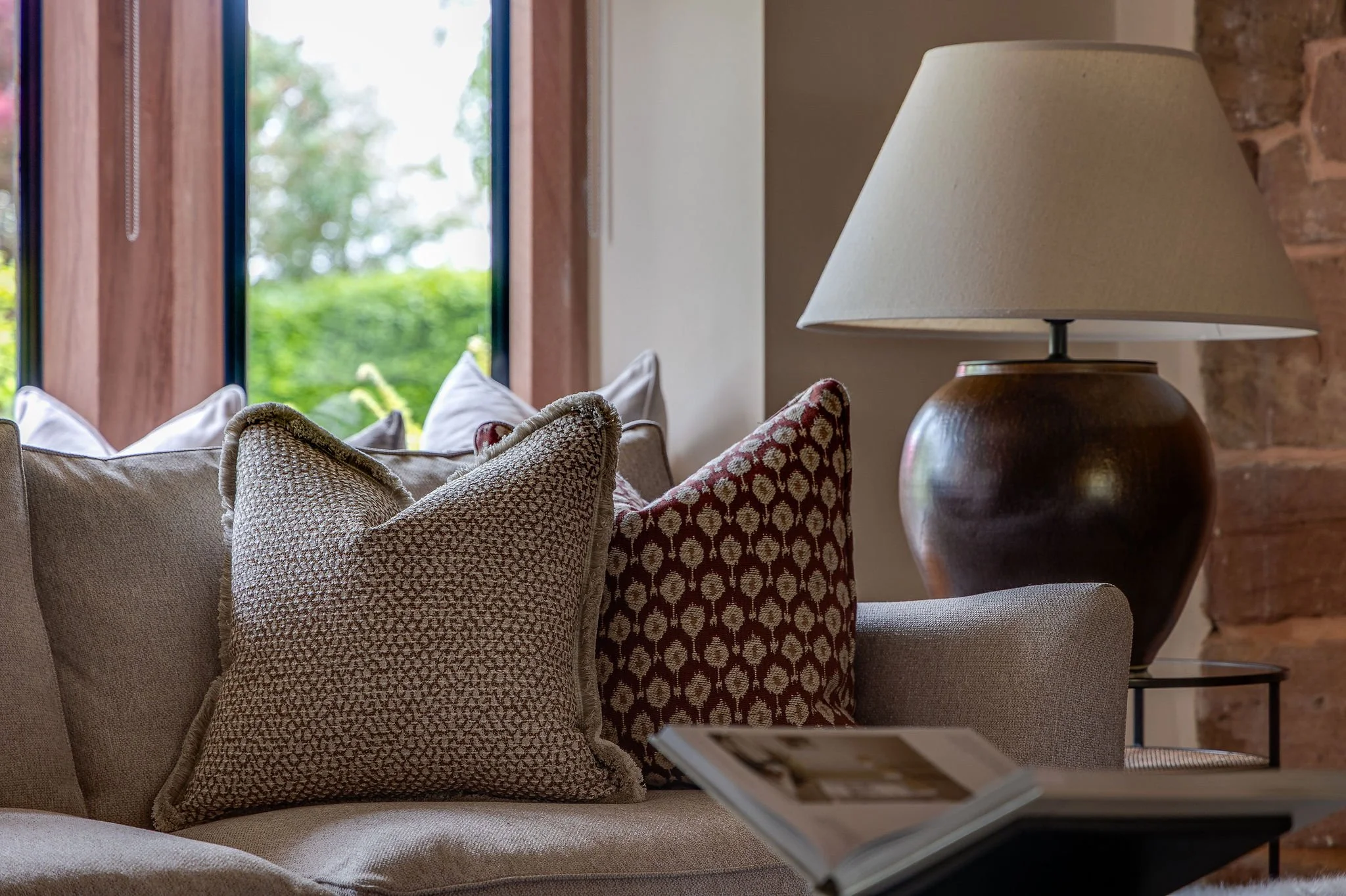 Close-up view of a beige sofa with textured and patterned cushions, a ceramic table lamp with a beige lampshade, and a window with blue trim showing green foliage outside in a cozy, rustic living room.