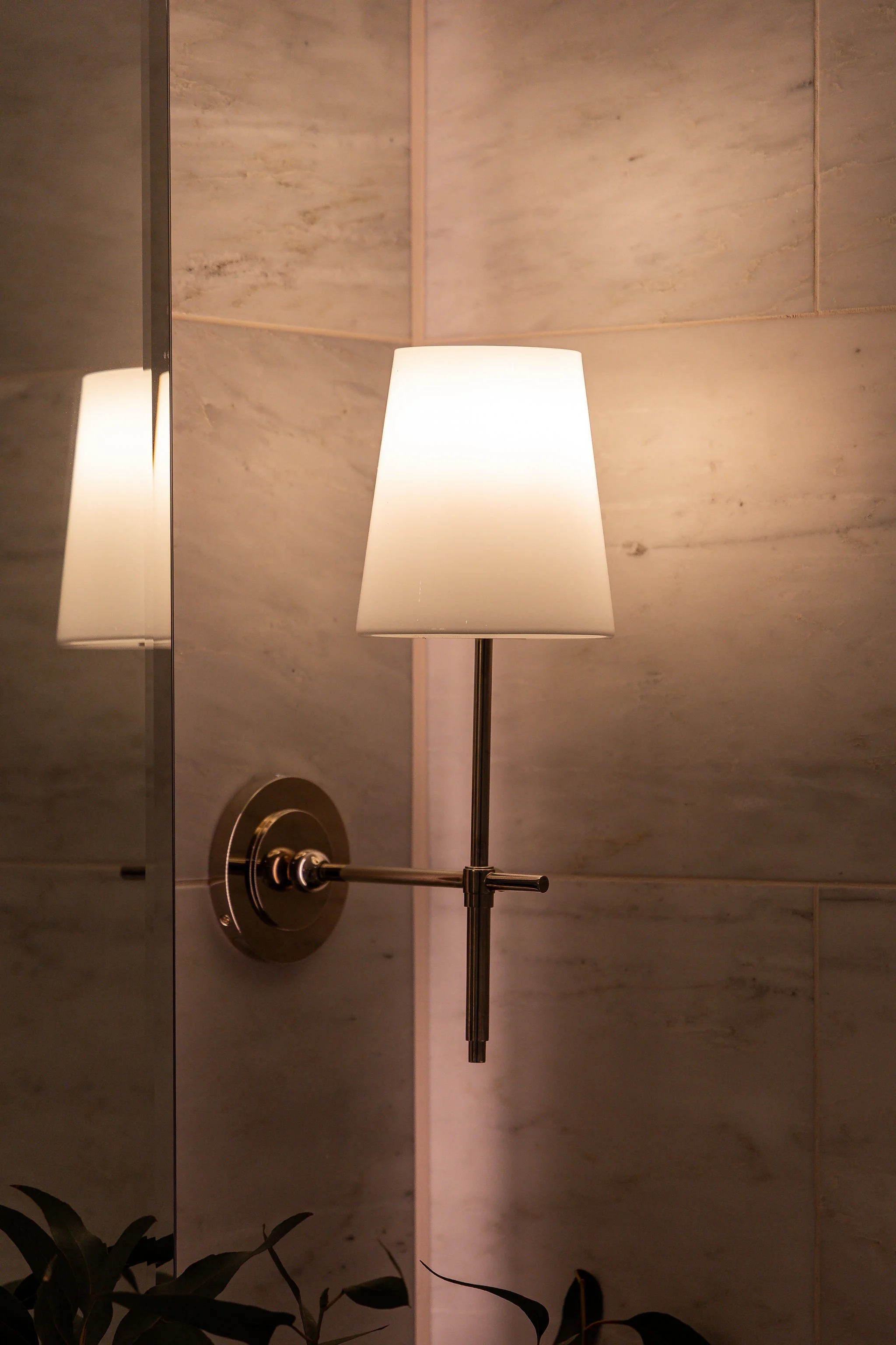 A modern wall-mounted lamp with a white lampshade in front of a beige tiled wall, reflected in a mirror.