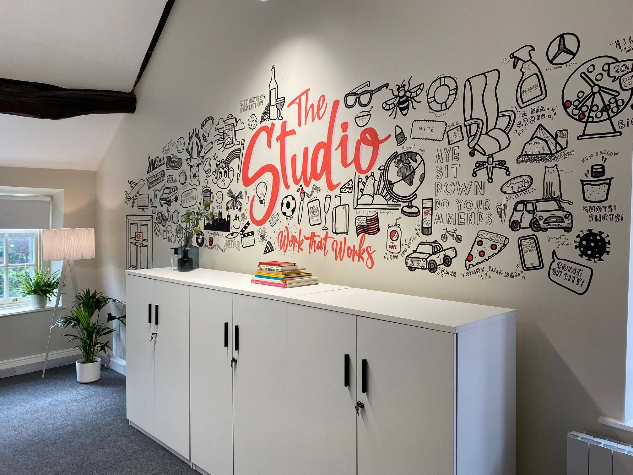 Interior of a room with a white storage cabinet, a window with a white shade, and a wall decorated with black and red doodles and text reading 'The Studio.' There are potted plants near the window and a small stack of books on the cabinet.