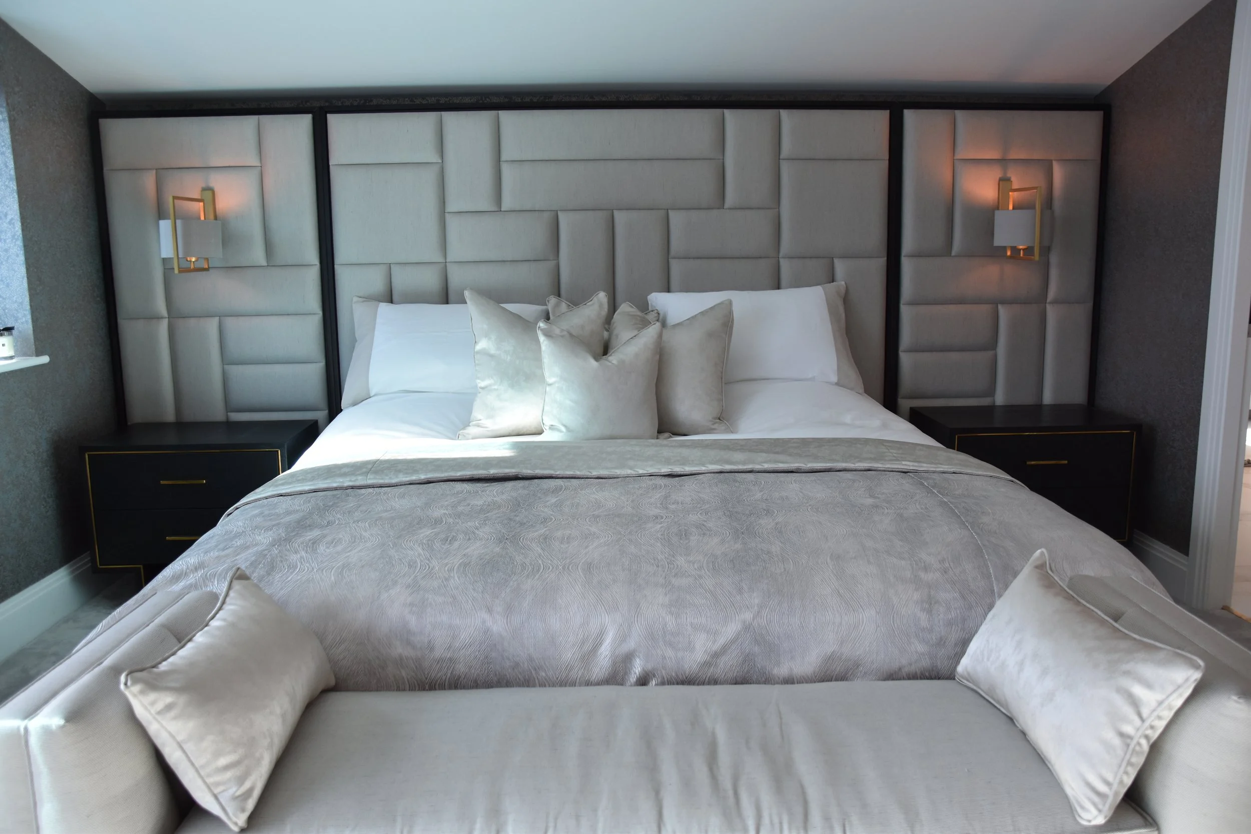 A neatly made bed with cream-colored pillows and bedding, set against an upholstered headboard with beige panels. Flanking the bed are two black nightstands with gold accents, each equipped with a wall-mounted light fixture. The room has soft, neutra