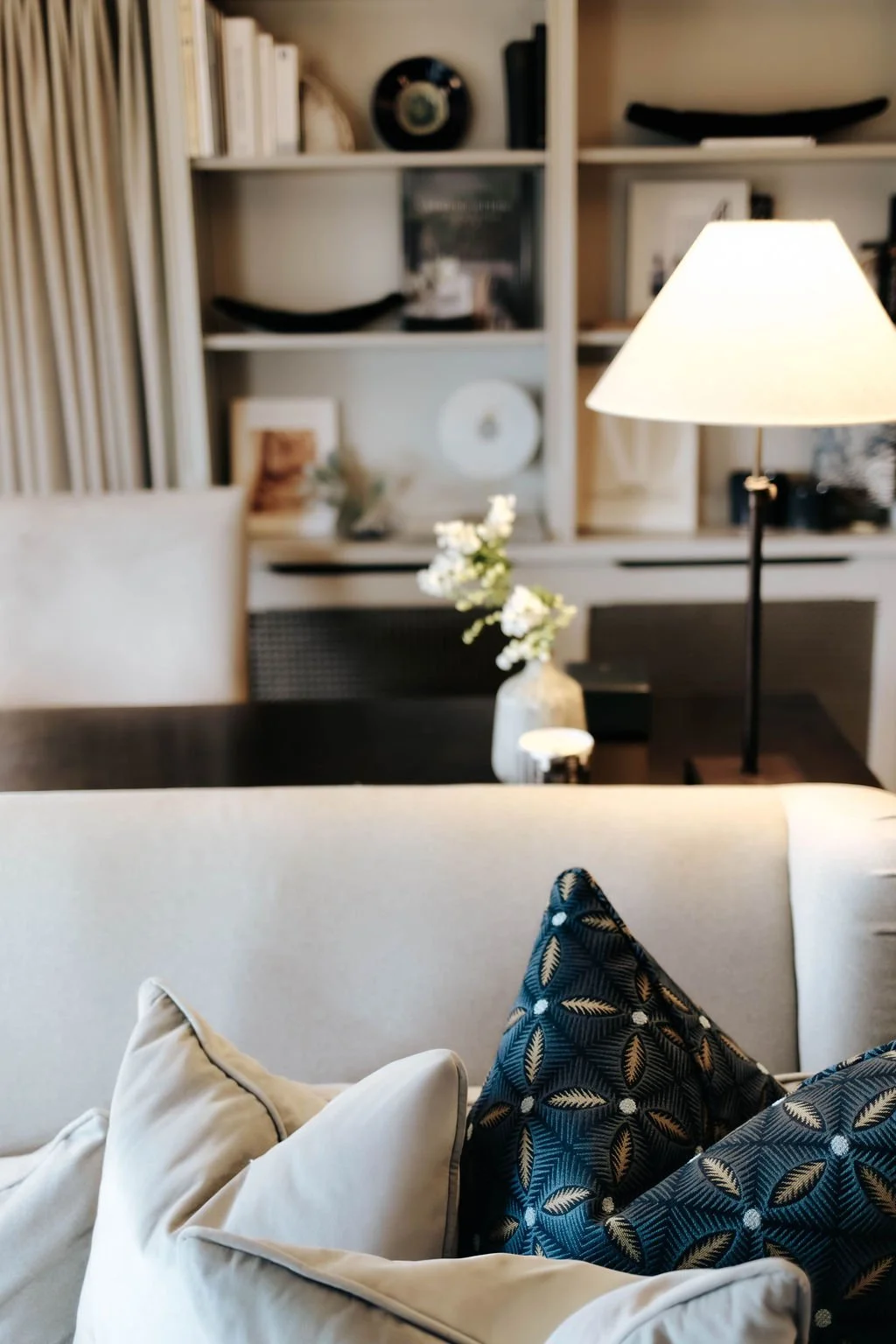 A cozy living room with a beige sofa and decorative pillows, a wooden coffee table with a vase of white flowers, a table lamp, and a white bookshelf in the background.
