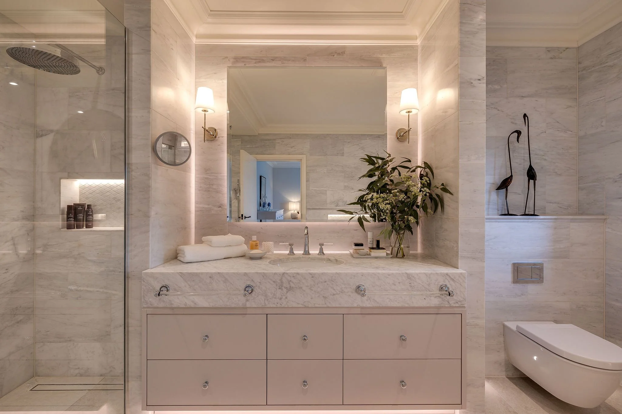 Luxury bathroom with marble walls and countertop, large mirror, two wall sconces, decorative sculpture of two birds, potted plant, and a toilet.