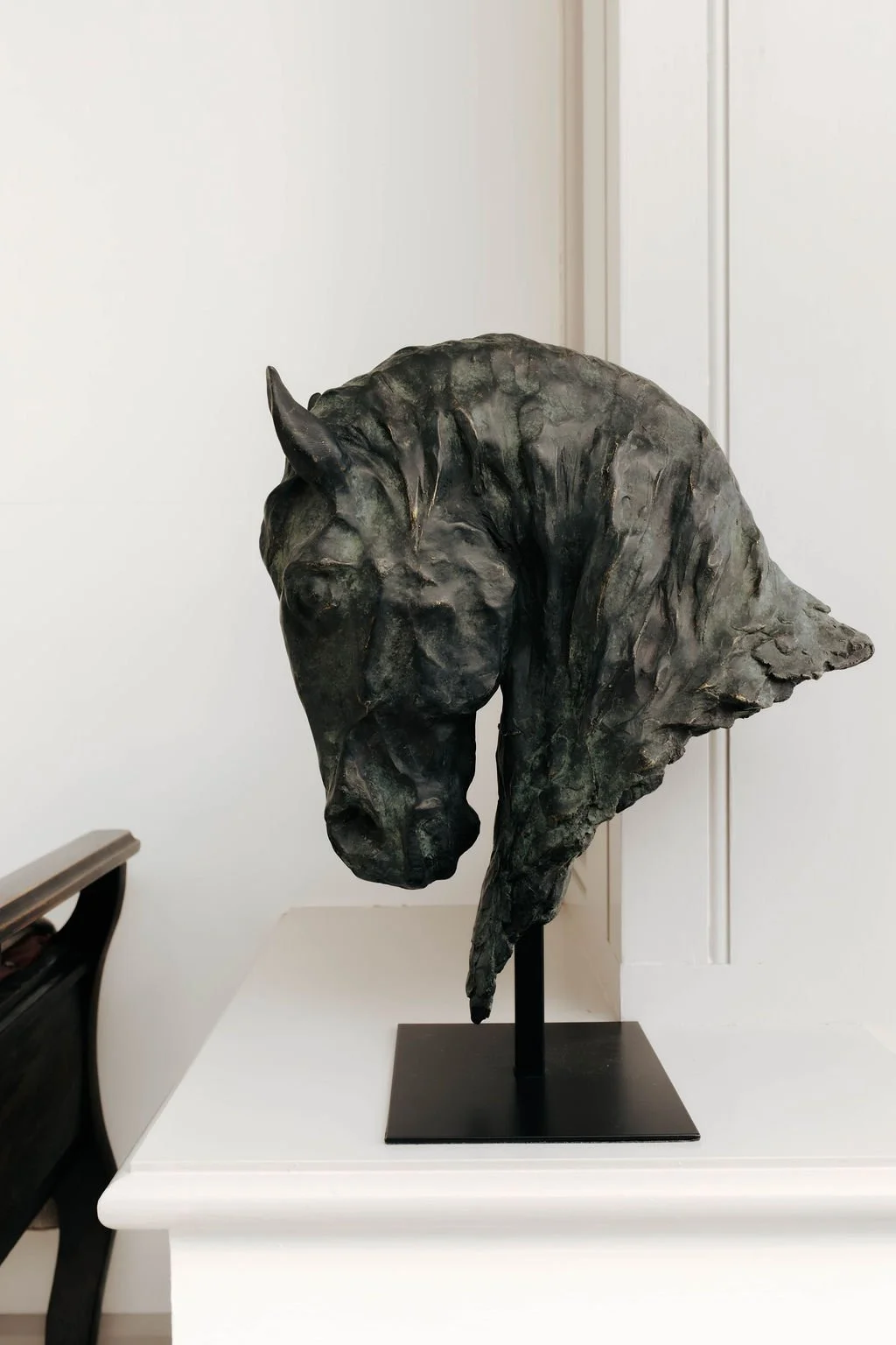 Bronze sculpture of a horse's head and part of its neck on a black stand, placed on a white table in a room with white walls.