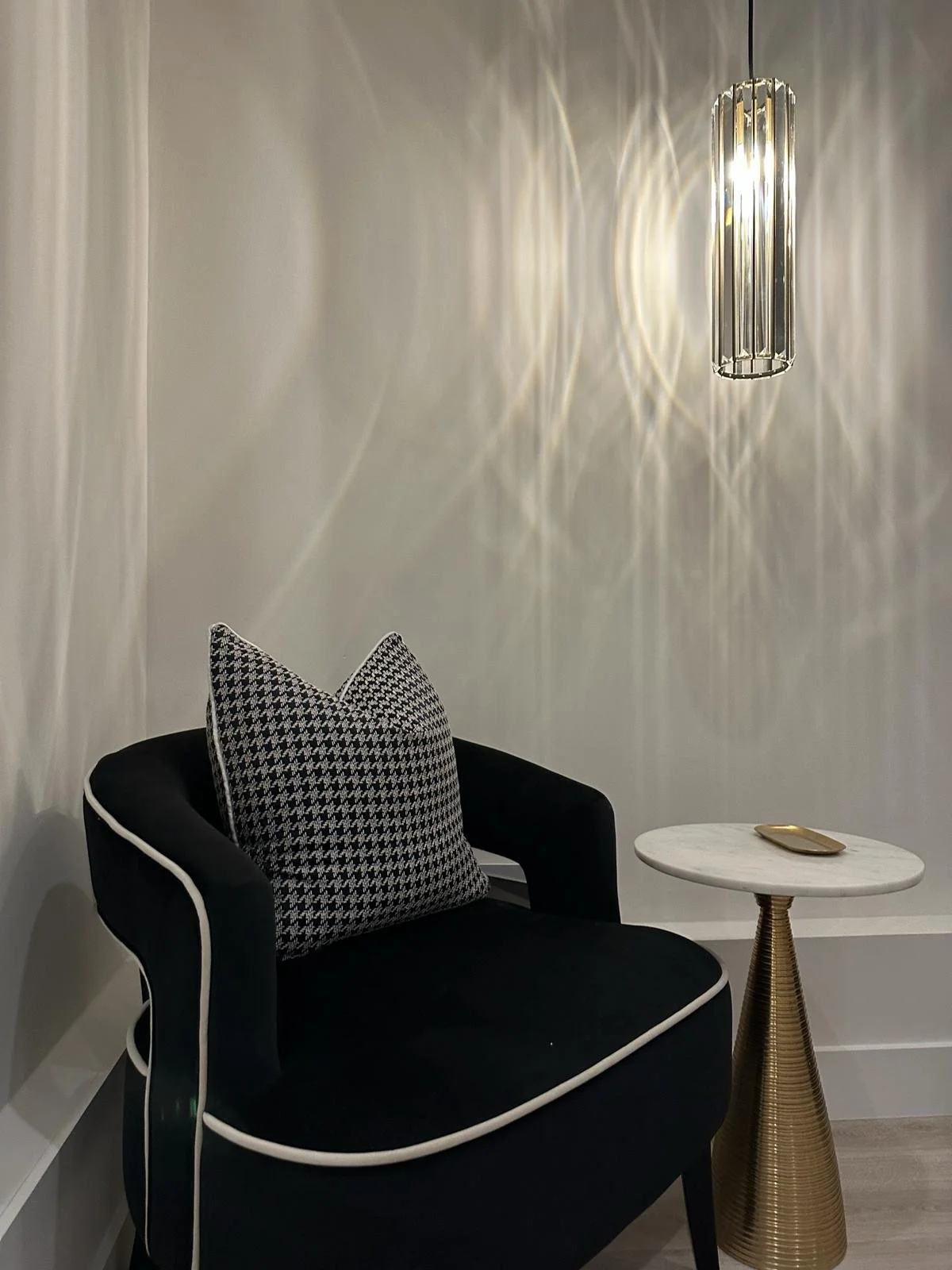 A black armchair with white piping and a black and white houndstooth patterned pillow, a small white marble side table with a gold decorative object, and a modern pendant light casting shadows on a white wall.