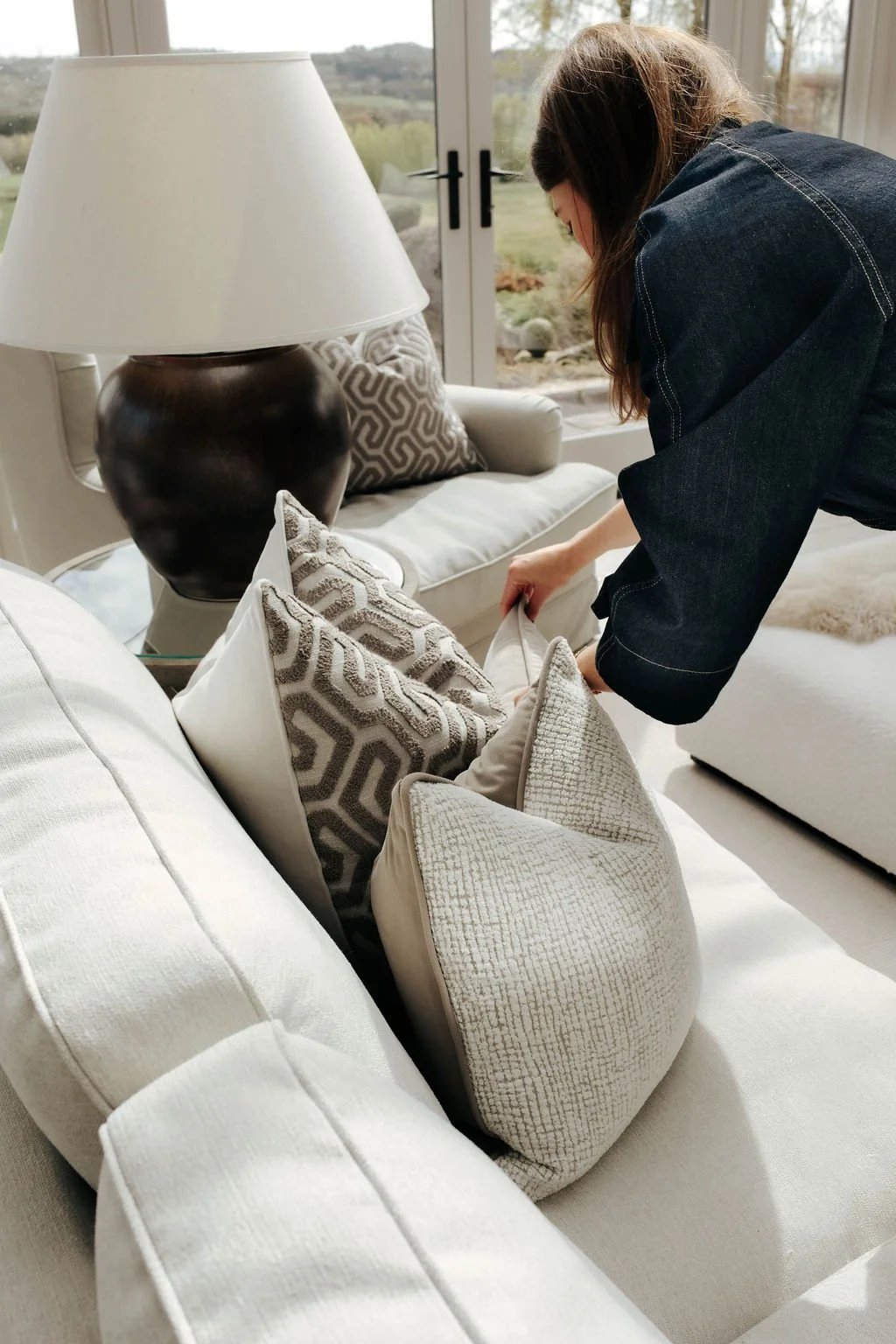 A woman arranges decorative pillows on a cream-colored sofa in a bright living room with large glass doors and a scenic view outside.