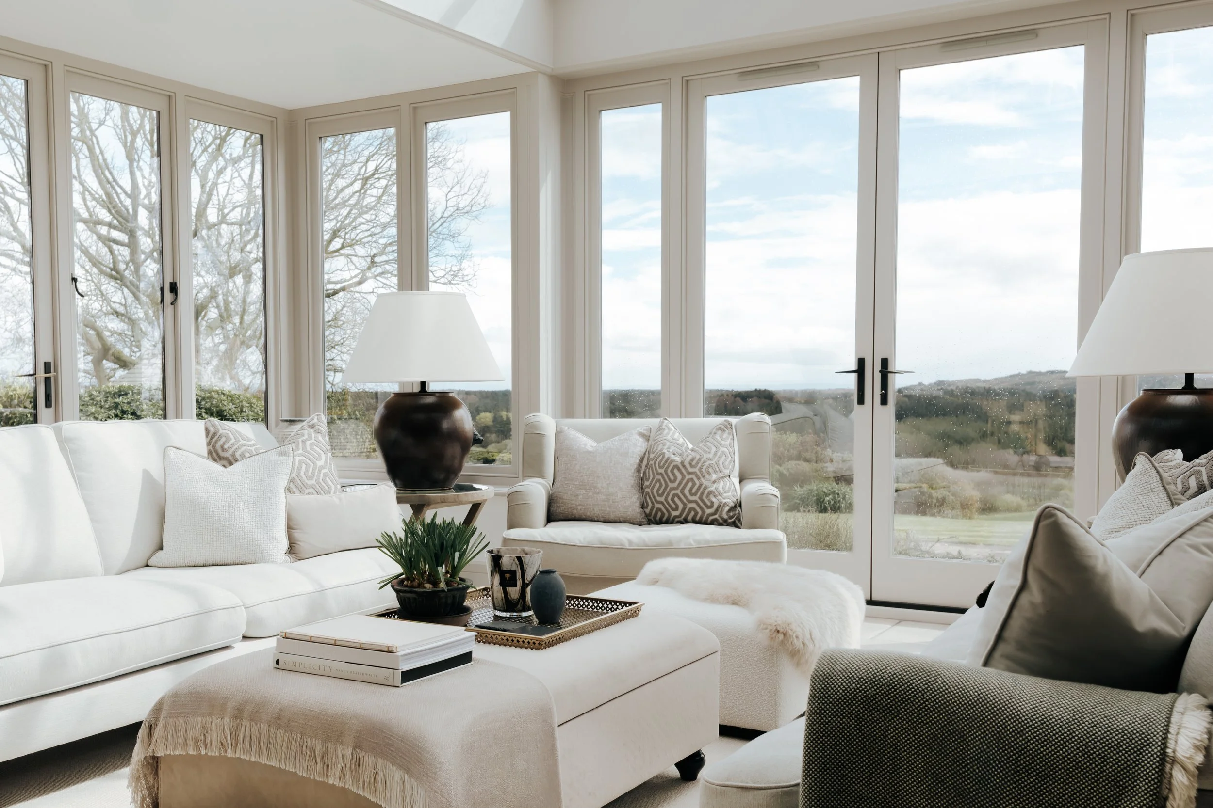 A bright living room with large windows showing a view of trees and sky outside. The room has white furniture, including a sofa with patterned throw pillows, and a coffee table with books, decorative vases, and a potted plant. Two table lamps are positioned on either side of the sofa.
