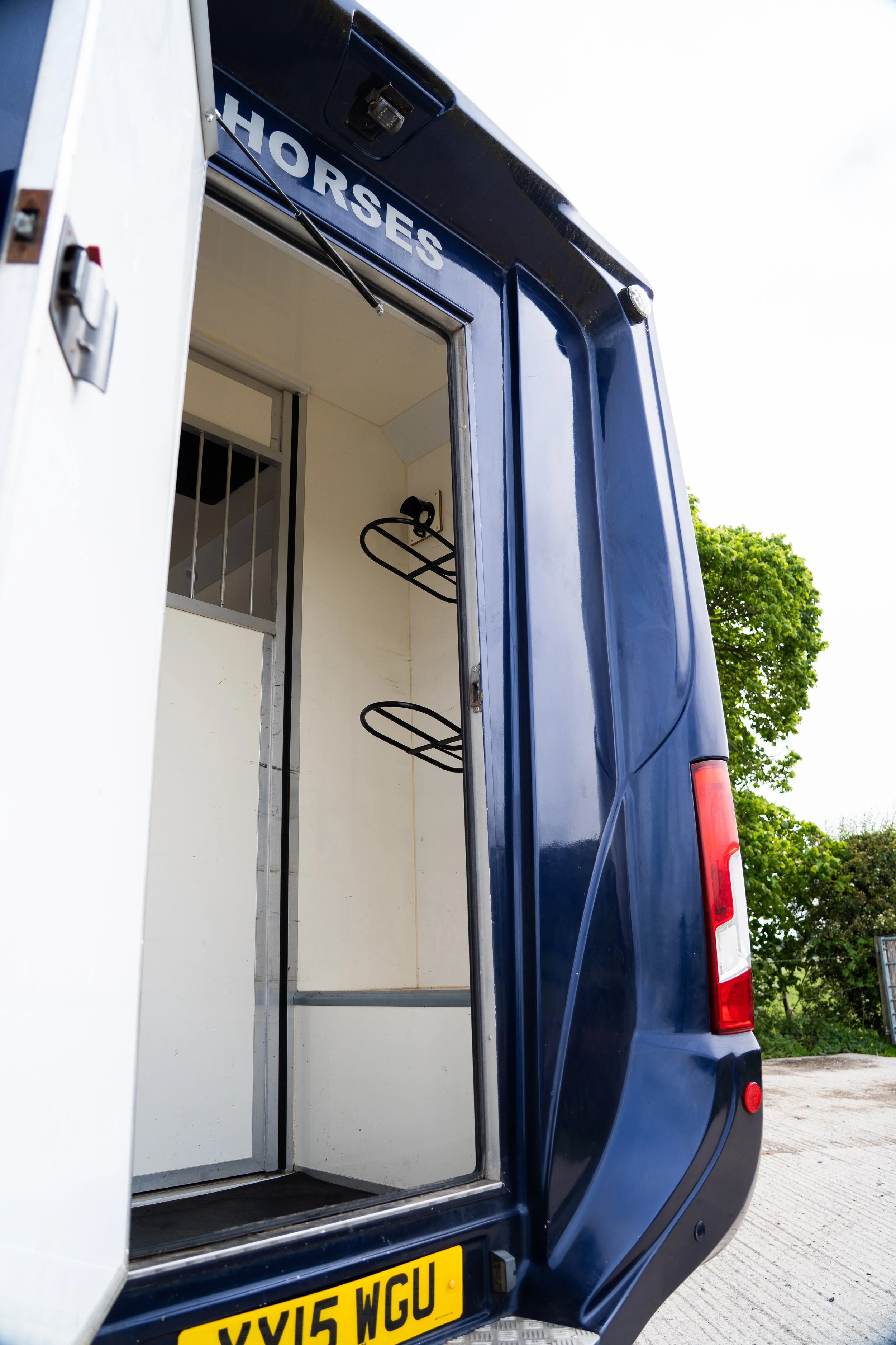 The back of a blue horsebox with the word 'HORSES' written on it. The horsebox door is open, showing the interior with metal bars and a grooming rack. A yellow license plate with the registration 'XYL5 WGU' is visible at the bottom. There are trees and a cloudy sky in the background.