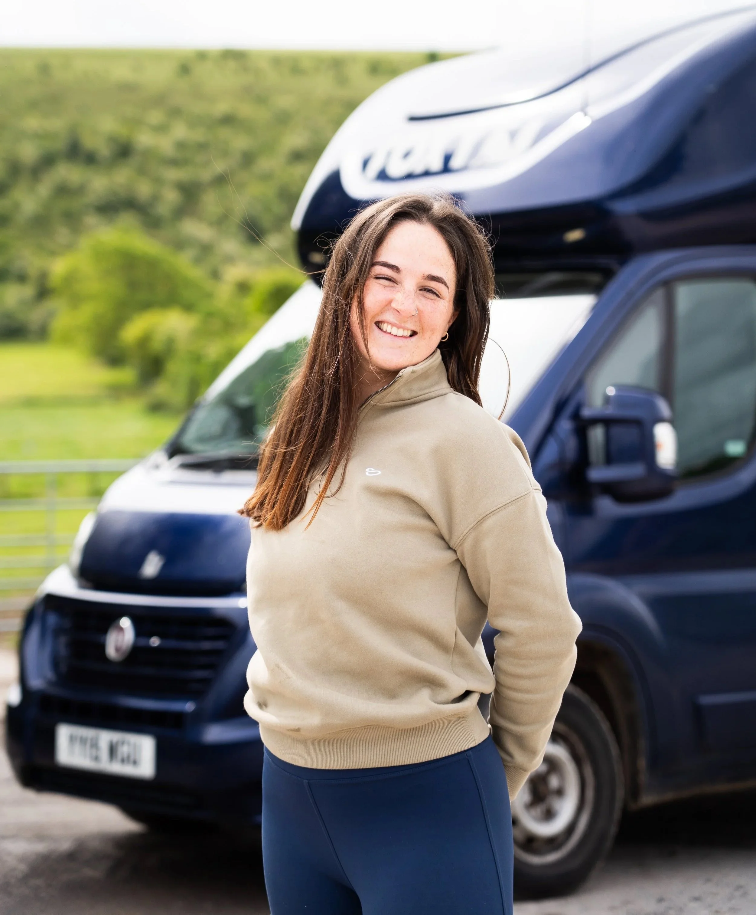 A young woman with long brown hair and a nose piercing is smiling and standing in front of a dark blue Volkswagen vehicle with a roof cargo box outdoors during daytime, with green trees and a grassy hillside in the background.