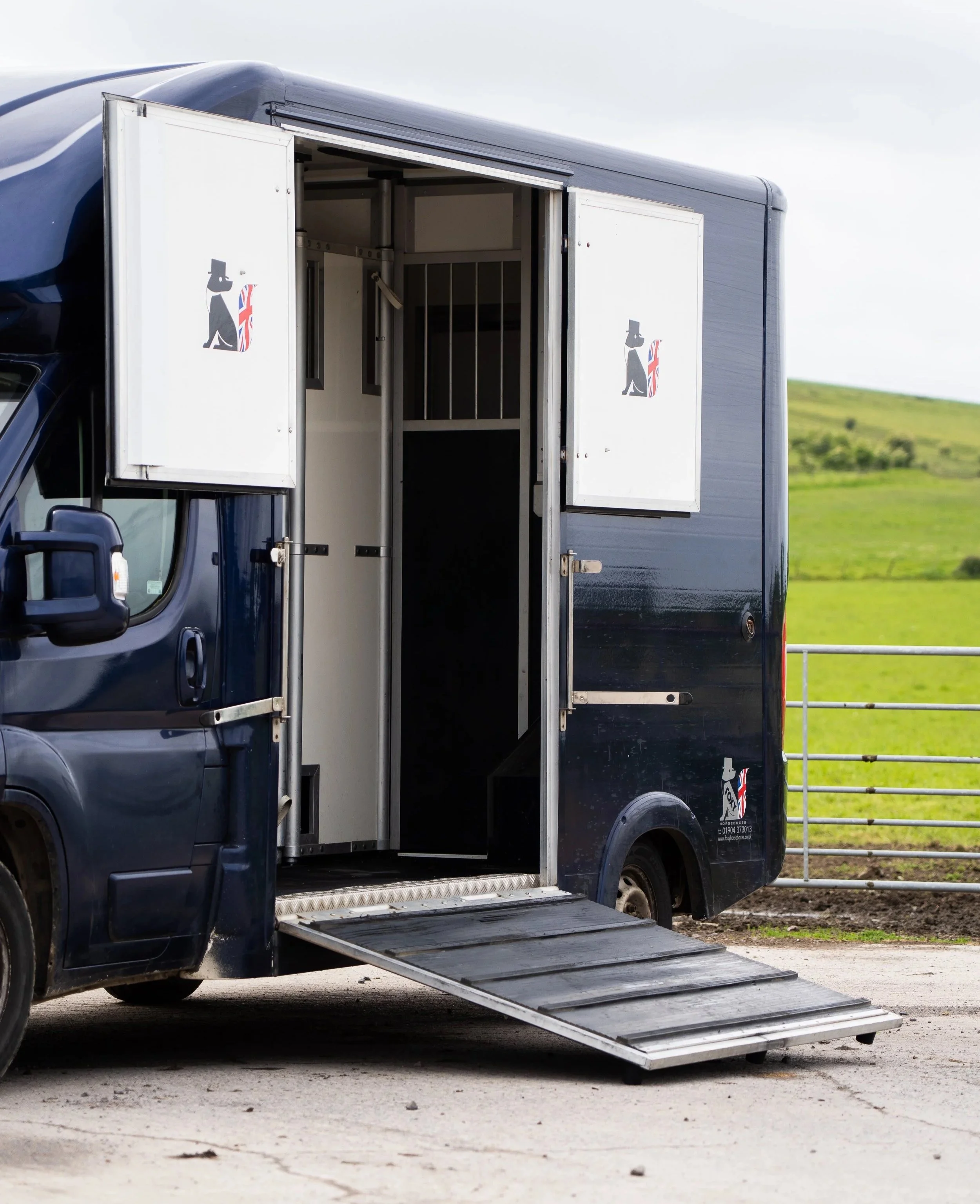 A black horse trailer with open doors, showing an interior with a partition, a ramp on the ground, and illustrations of a dog and a horse with Union Jack flag designs on the sides. The trailer is parked on a paved area with green fields in the background.