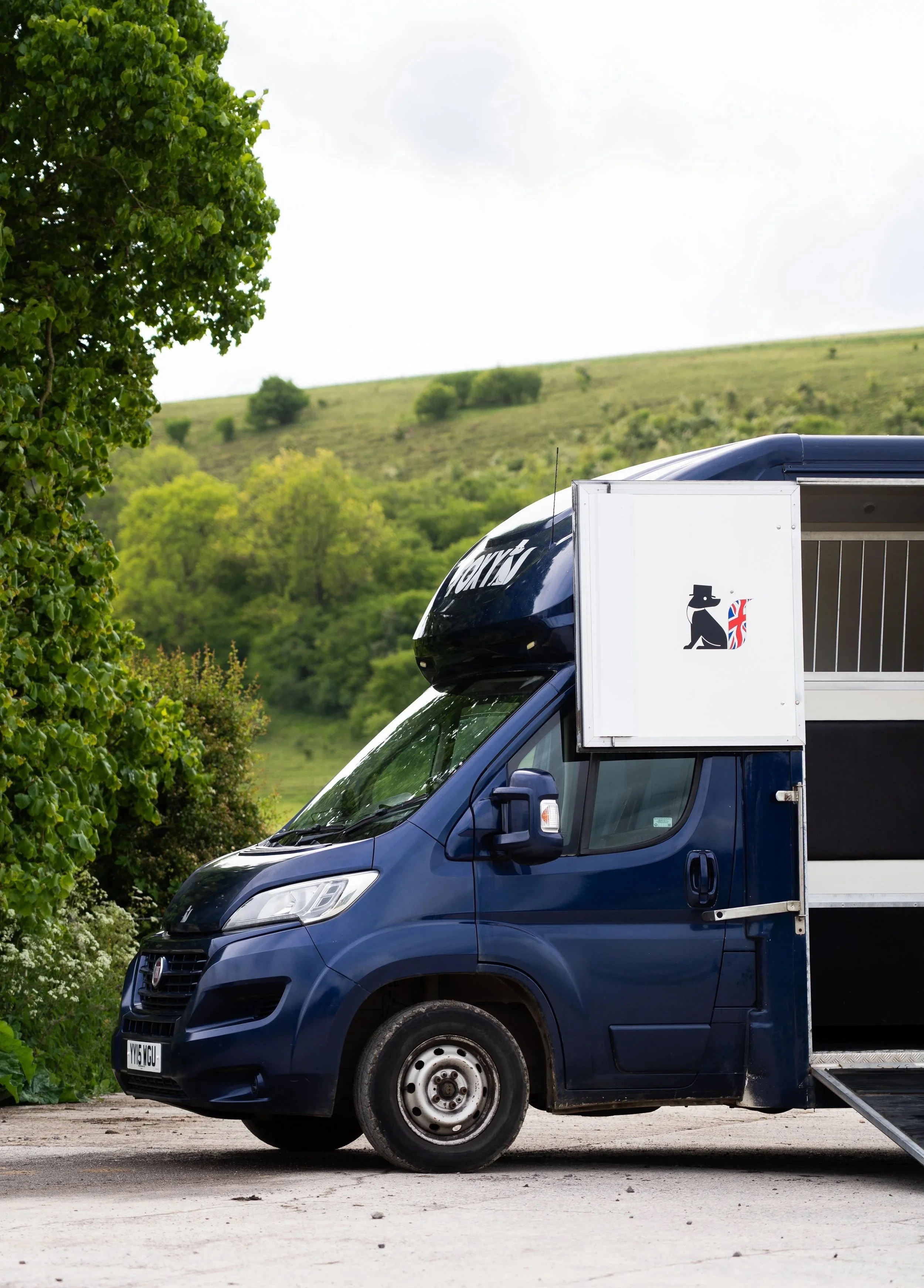 A black and white delivery truck with a dog logo on the side, parked on a gravel surface with greenery and a hill in the background.
