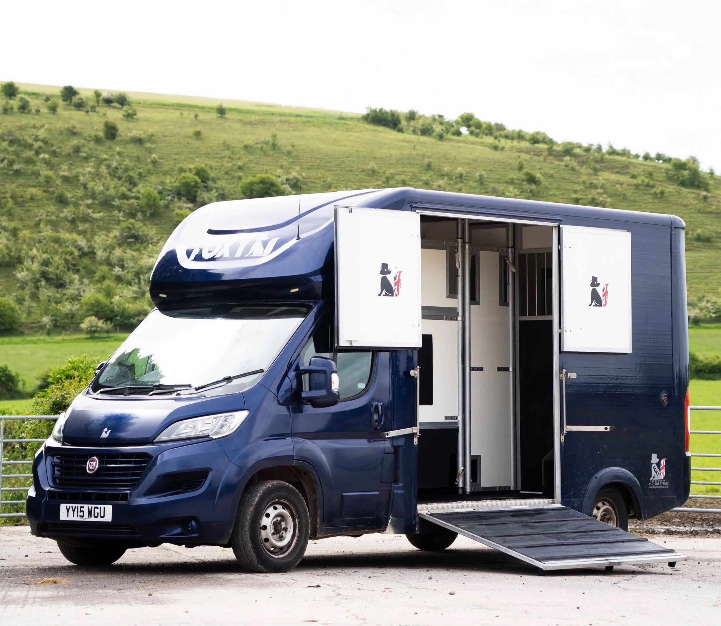 A blue Fiat delivery van with an open side door, parked on a paved area with a scenic green hillside in the background.
