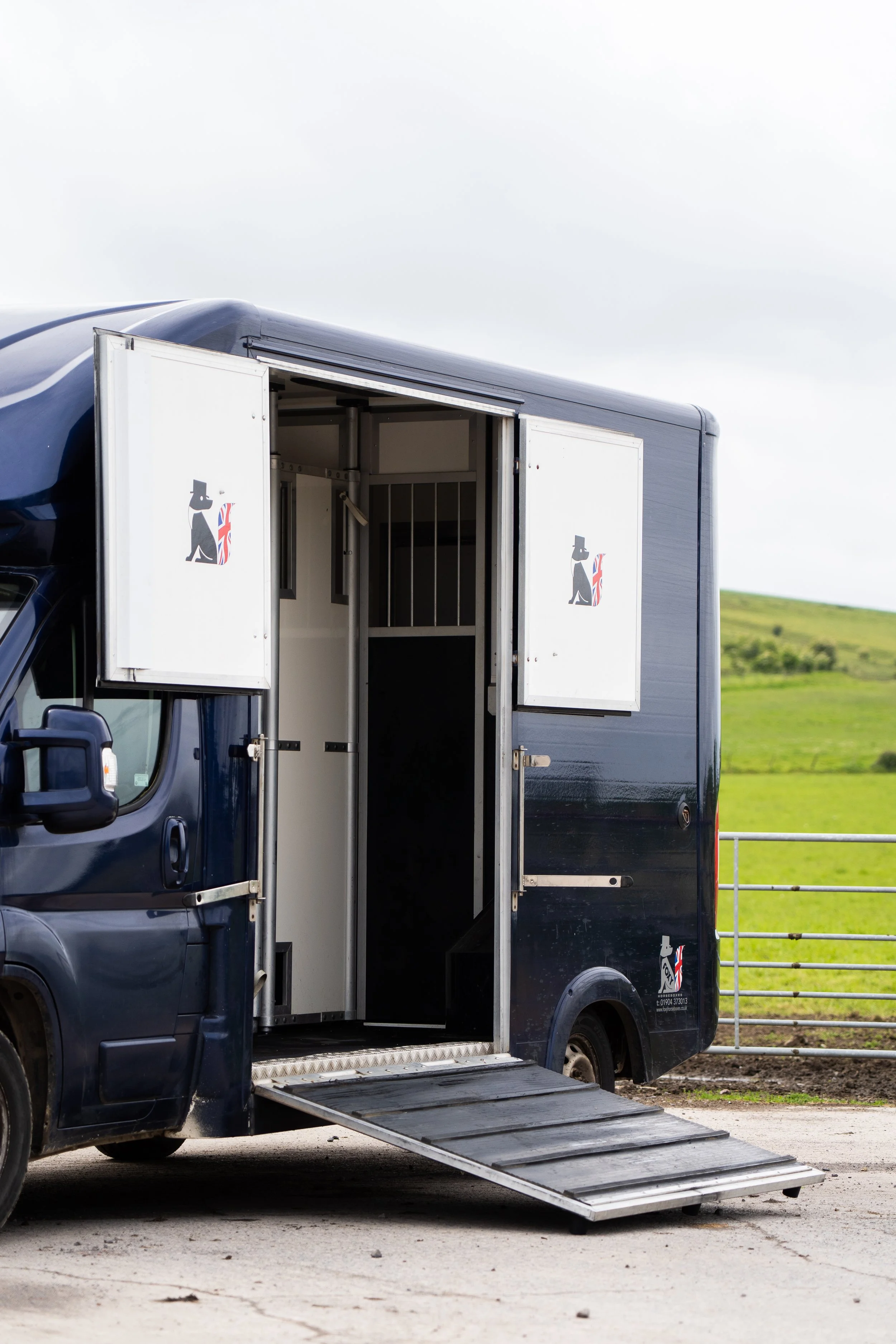 A black pet transport truck with a lowered metal ramp extended on the ground outside, showing an open side door with a dog logo, set in a rural area with green hills in the background.