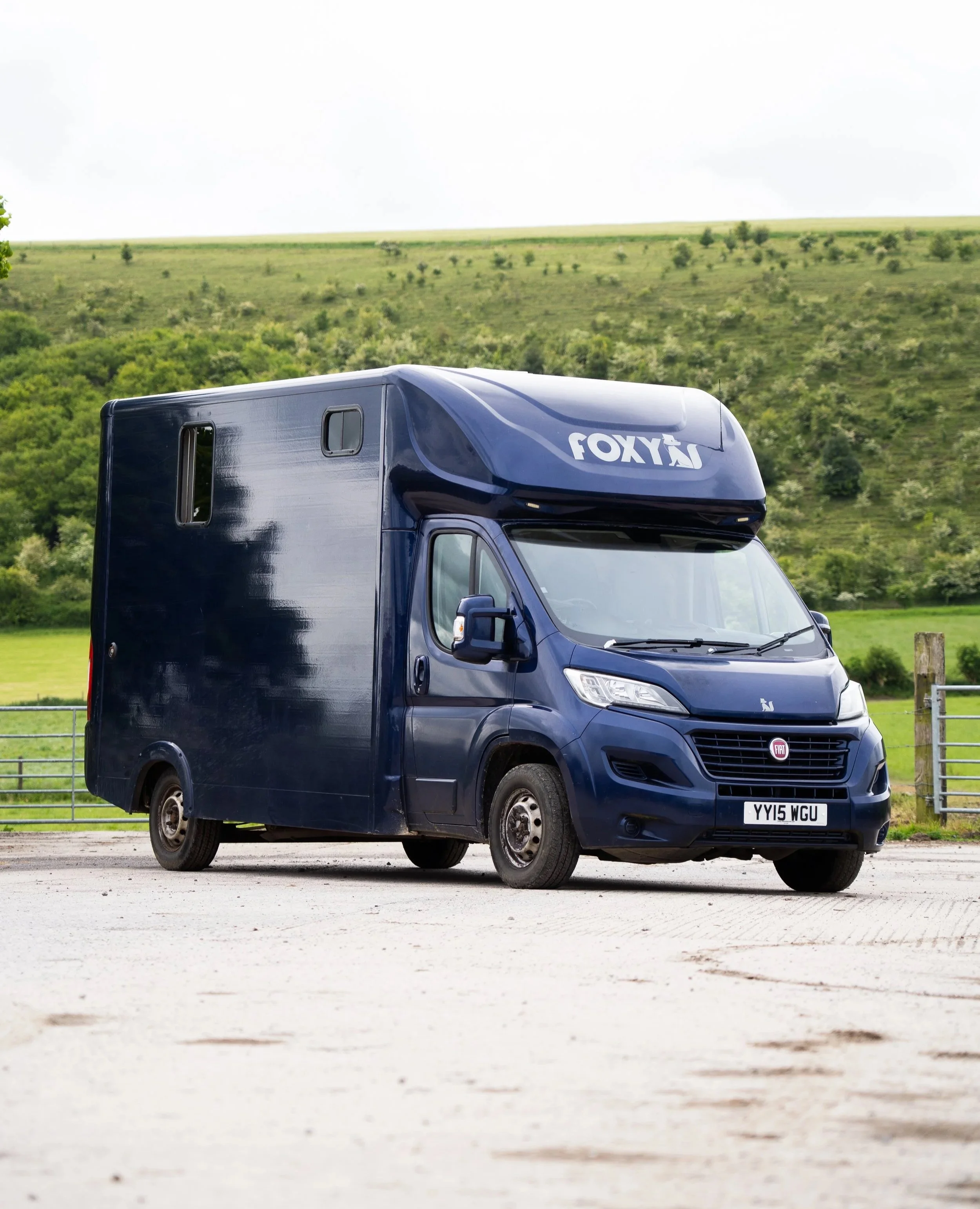 A dark blue camper van parked on a gravel surface with green hills and trees in the background.