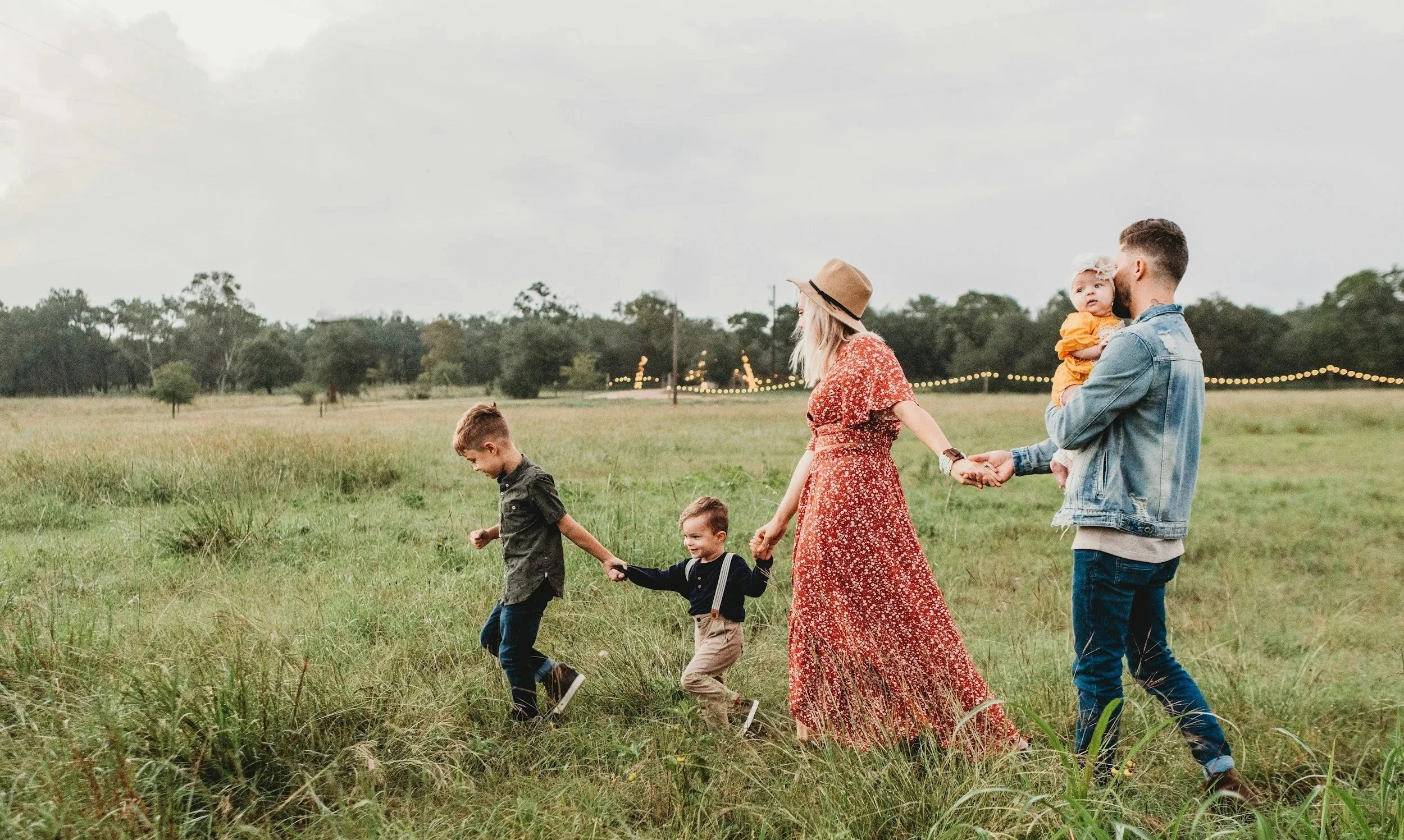 A family of five walking through a grassy field outdoors during daytime, including a man, woman, three children, and a baby in the man's arms, with trees and string lights in the background.