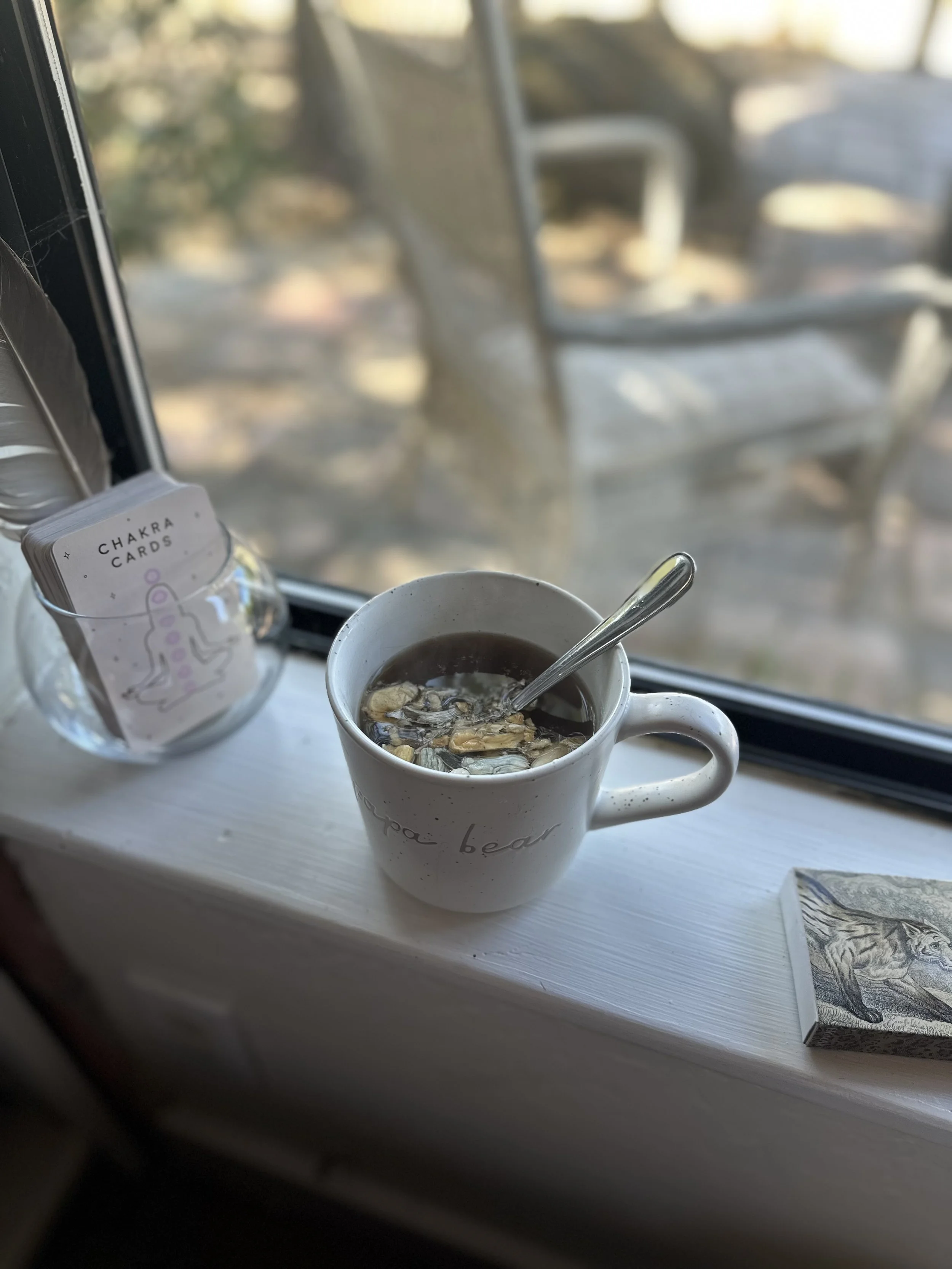 A white mug filled with tea sits on a windowsill, with a spoon inside. To the left of the mug, there is a glass jar of chakra cards. Outside the window, there are blurred outdoor chairs and a table, with sunlight filtering through.