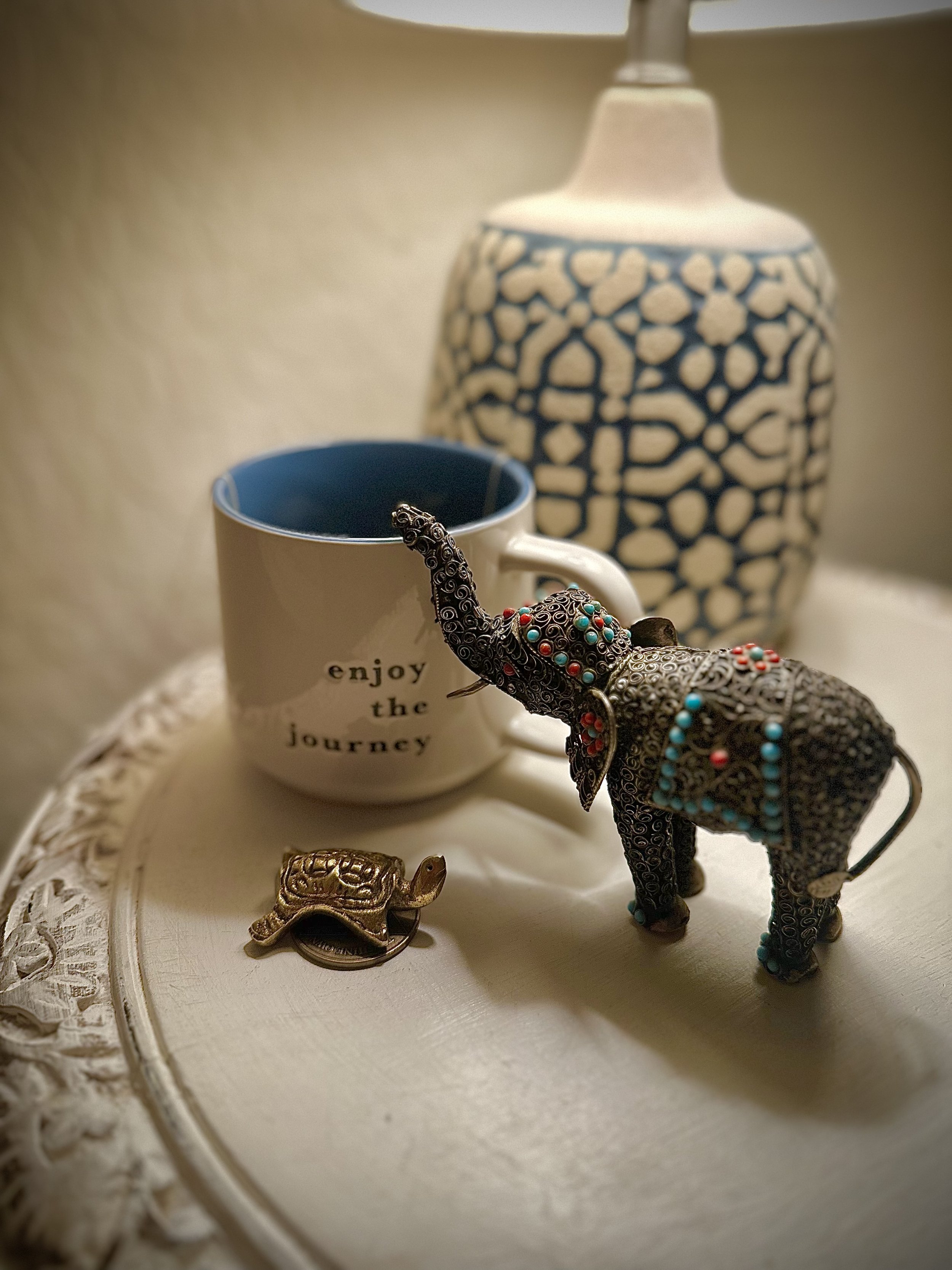 A decorative elephant figurine decorated with turquoise and red beads, with a smaller turtle ornament nearby, on a vintage tray with intricate lace trim, alongside a white mug with the words 'enjoy the journey,' and a blue and cream patterned vase in the background.