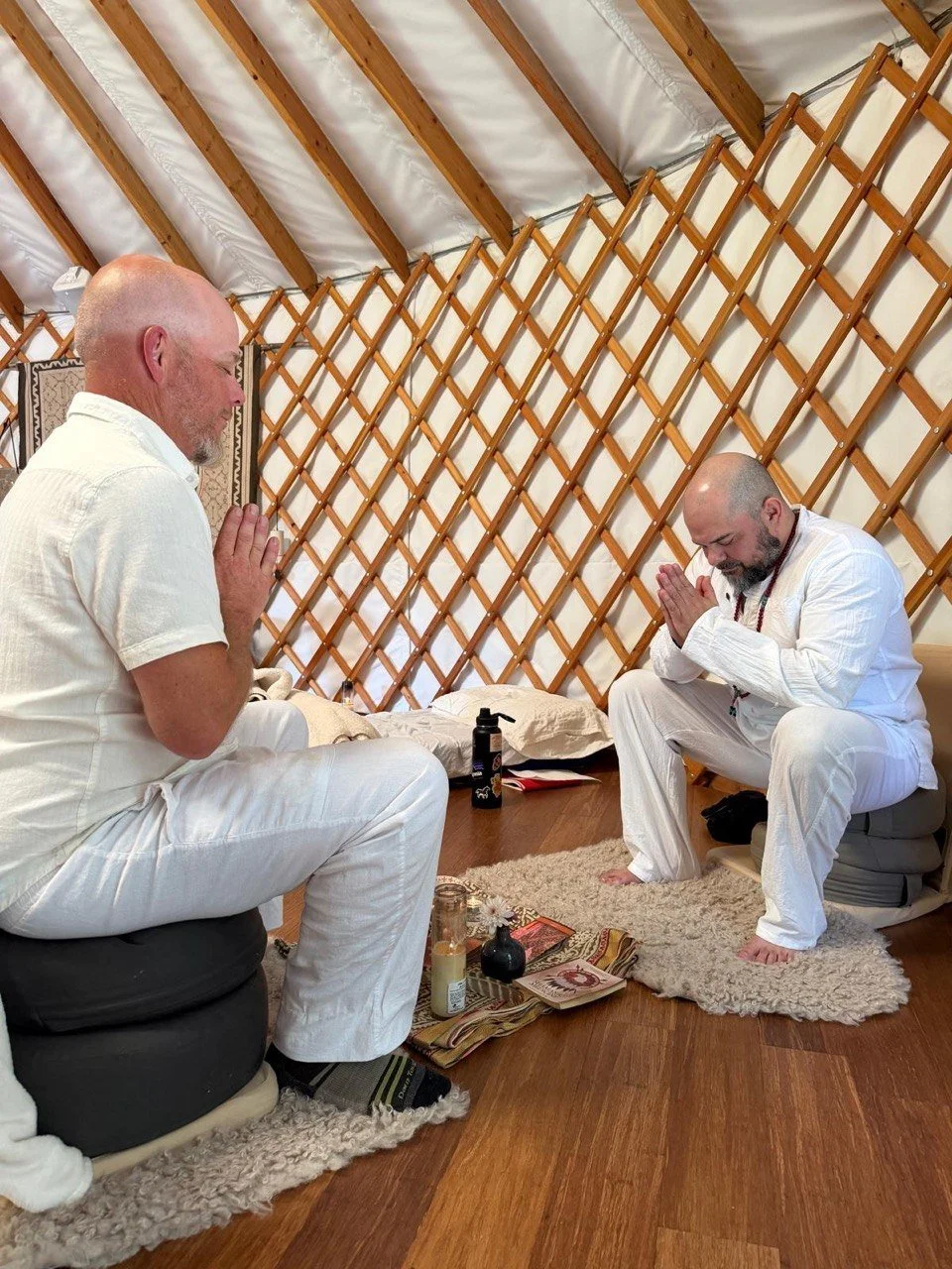 Two men with shaved heads and beards dressed in white clothing sit on cushions in a yurt, praying with hands pressed together and heads bowed. They sit on shaggy rugs with spiritual objects and candles placed on a mat between them.