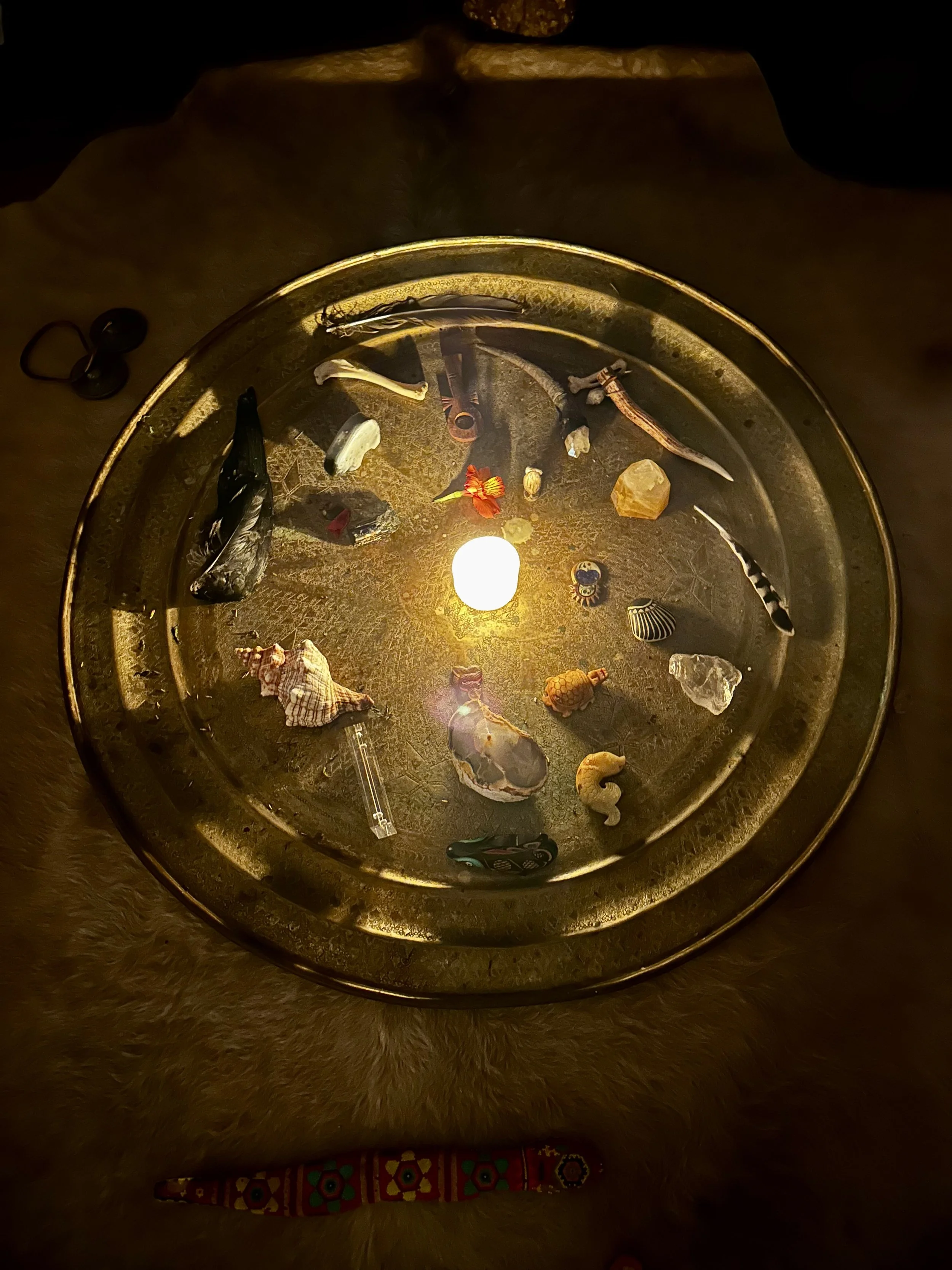 A round decorative tray with various small objects including shells, a candle, a feather, small figurines, and stones, illuminated by a single warm light at the center.