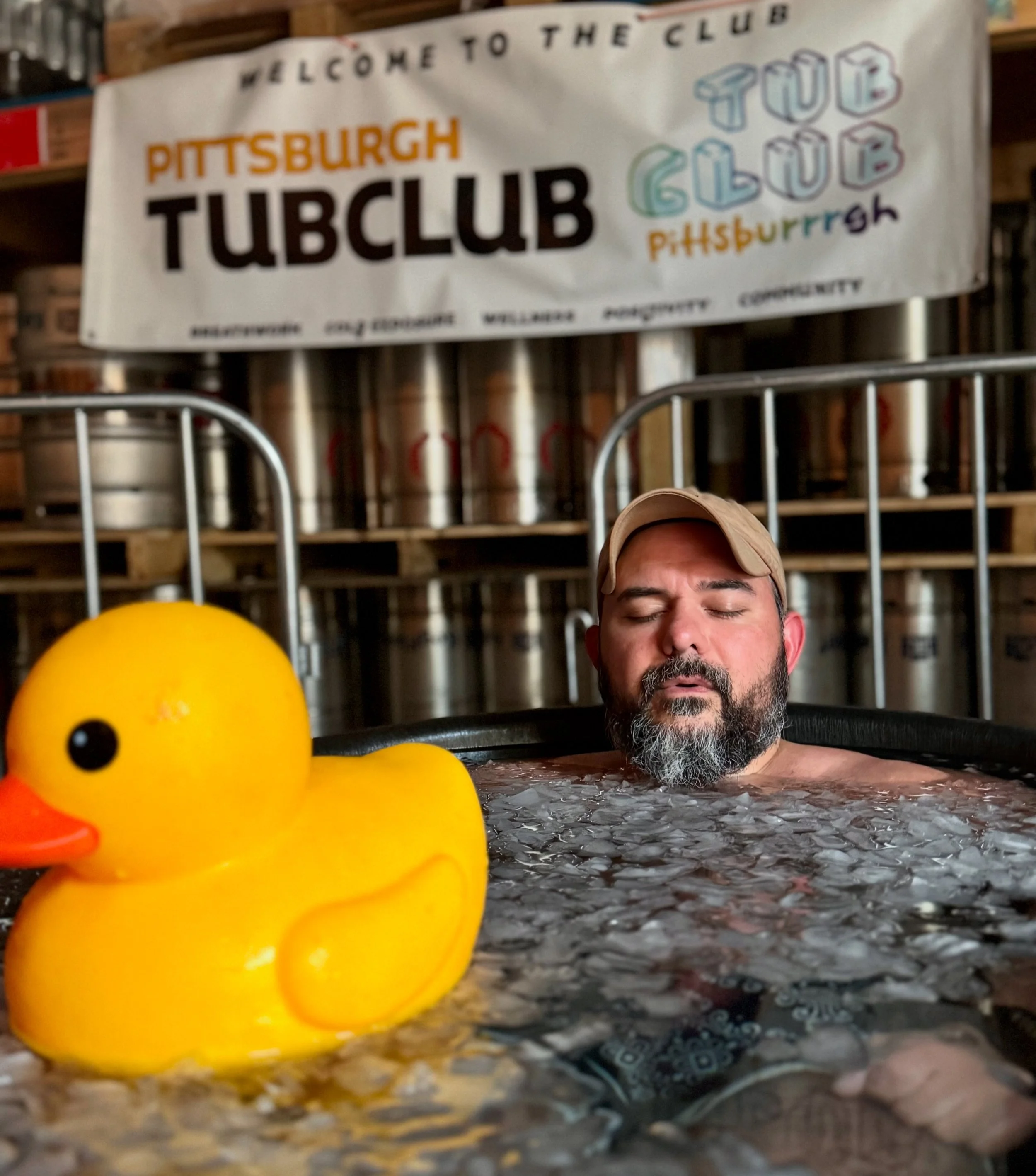 Close-up of a rubber duck on a wet surface with a man's face in the background and a banner that reads 'Welcome to the club Pittsburgh Tubclub'.