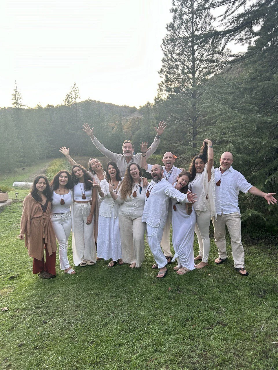 Group of people outdoors on a grassy field, dressed in white and beige clothing, smiling and celebrating with joyful poses, trees in the background.