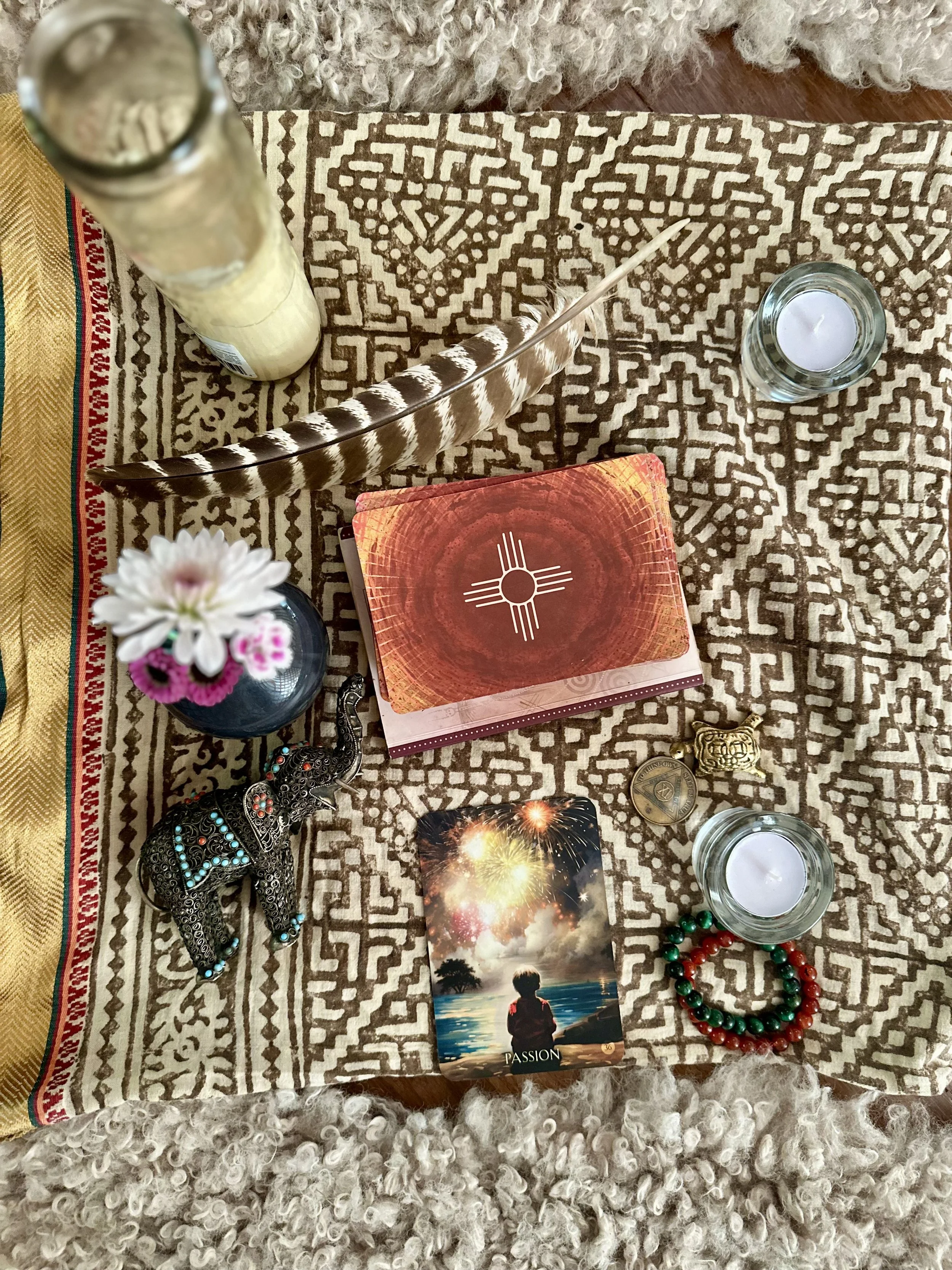 Decorative table with spiritual items including a feather, candles, a floral arrangement, a small elephant figurine, a dreamcatcher, a tarot card titled "Passion" with a boy by the water, a bracelet of red and green beads, and a chest of drawers.