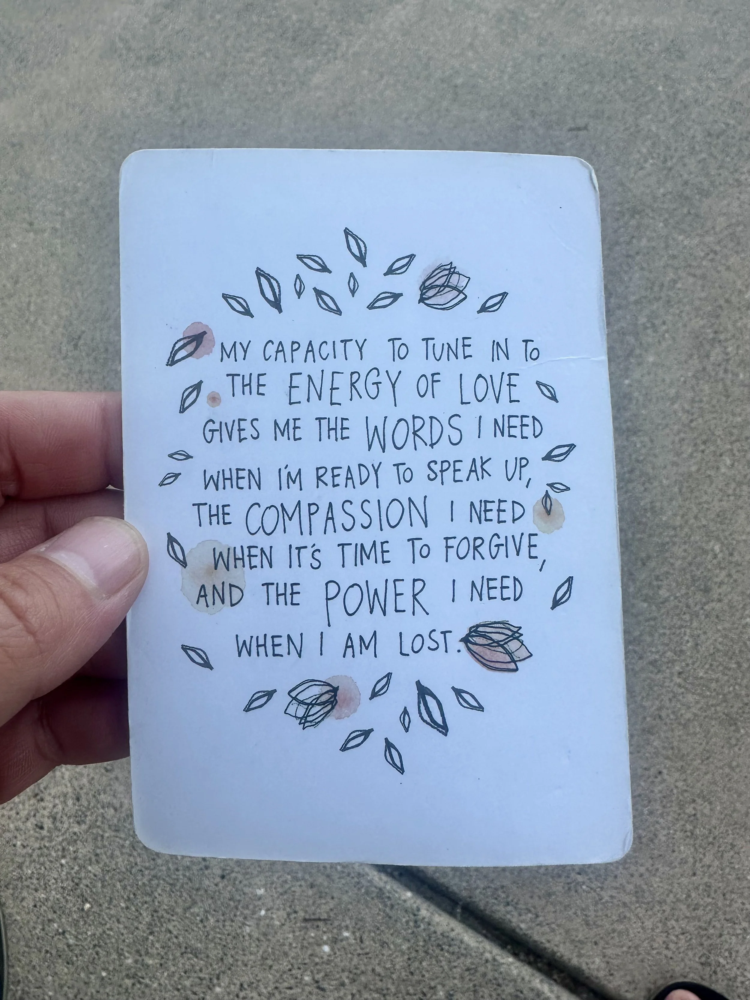 A handwritten note on a piece of paper held by a person's hand, featuring a quote about love, forgiveness, and inner strength, decorated with small hand-drawn leaves and abstract shapes.
