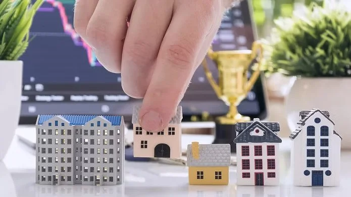 A person's hand places a house-shaped object among other miniature houses on a desk with a laptop showing stock charts in the background.
