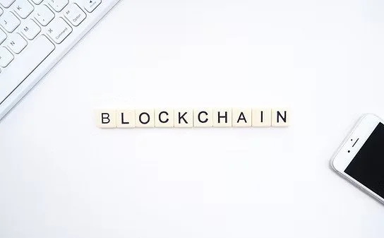 Scrabble tiles spelling 'BLOCKCHAIN' on a white surface, with a keyboard partially visible in the top left corner and a smartphone in the bottom right corner.