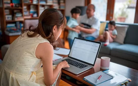 Woman working on a laptop at a desk with children in the background