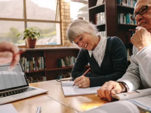 Two elderly people laughing and sitting at a table in a library or study room, with bookshelves and a window in the background.