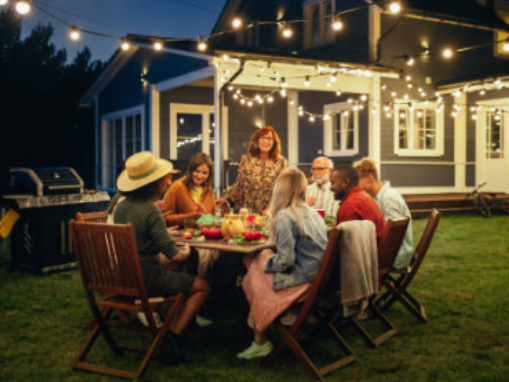 A group of people having a dinner party outdoors at night with string lights overhead, sitting around a table on a lawn in front of a house.