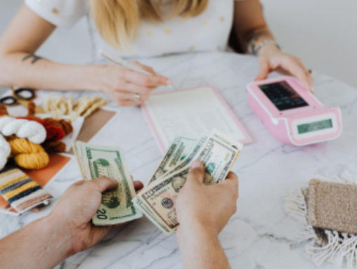 Person counting money at a table with knitting supplies and a pink calculator nearby.
