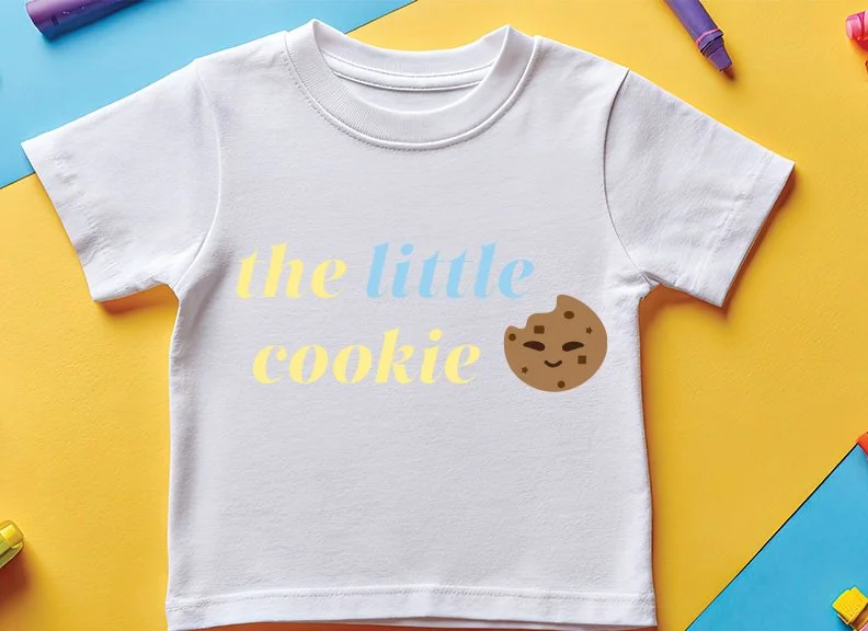 White children's t-shirt with yellow and blue text that reads "the little cookie" and a smiling cookie illustration, placed on a yellow background with colored chalk around it.