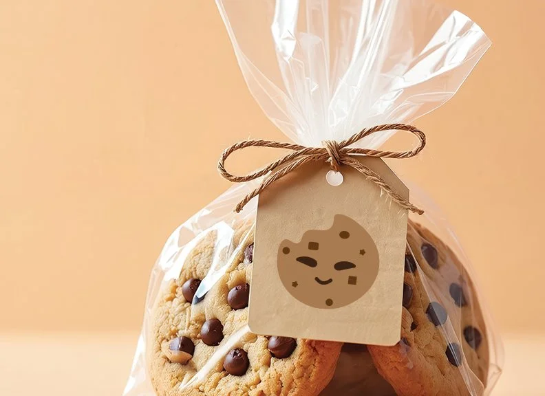Cookies with chocolate chips in a clear plastic bag tied with twine, with a gift tag featuring a smiling cartoon bear face printed on it.