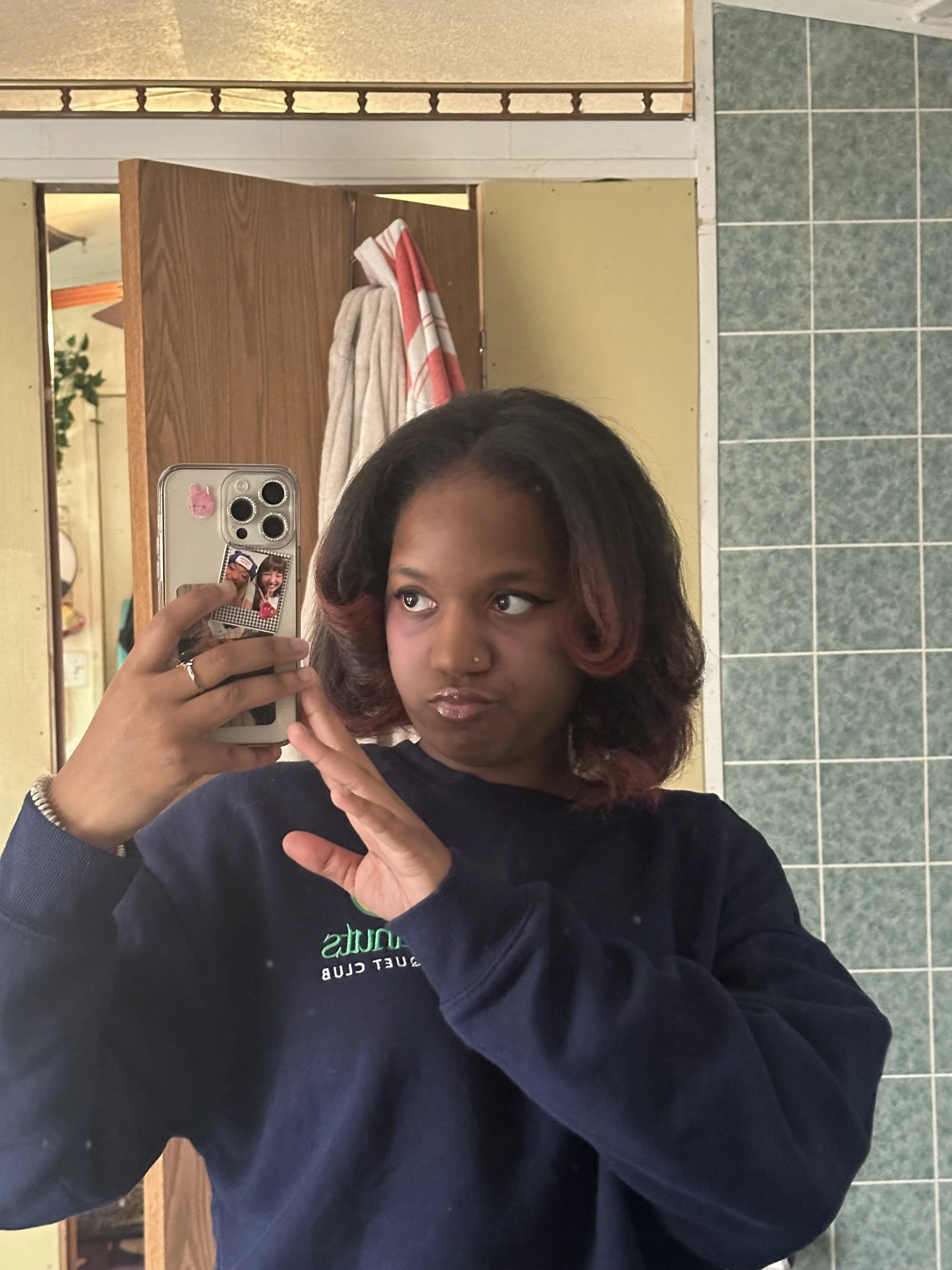 A person with shoulder-length dark hair with pink highlights taking a mirror selfie in a bathroom. They are wearing a black long-sleeve shirt with some green and white text. The bathroom has a green tiled wall and a wooden counter with towels and a mirror visible in the background.