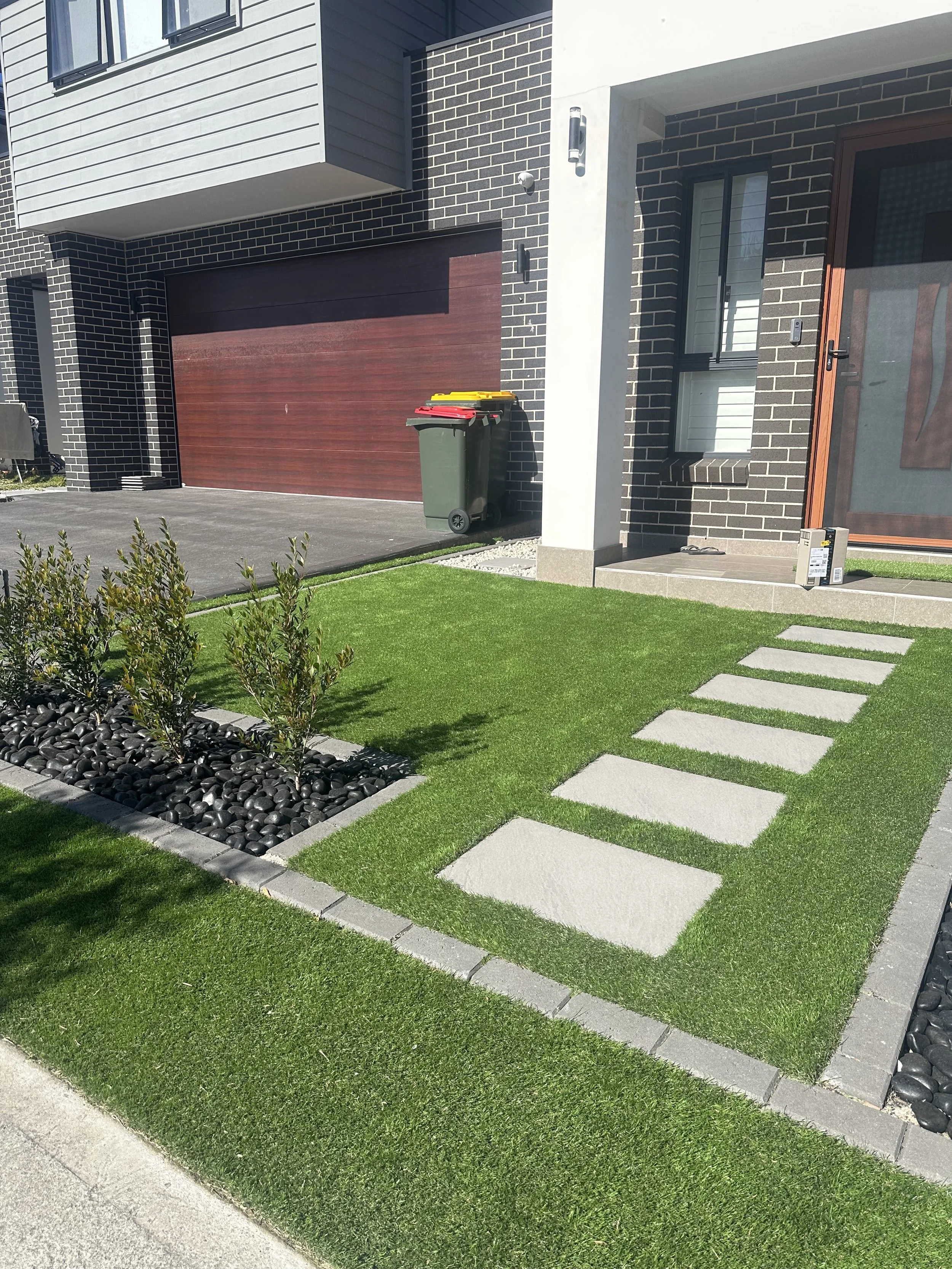 Synthetic grass for small garden bed with bushes and black decorative rocks - Services by Greenscape Turfing Sydney